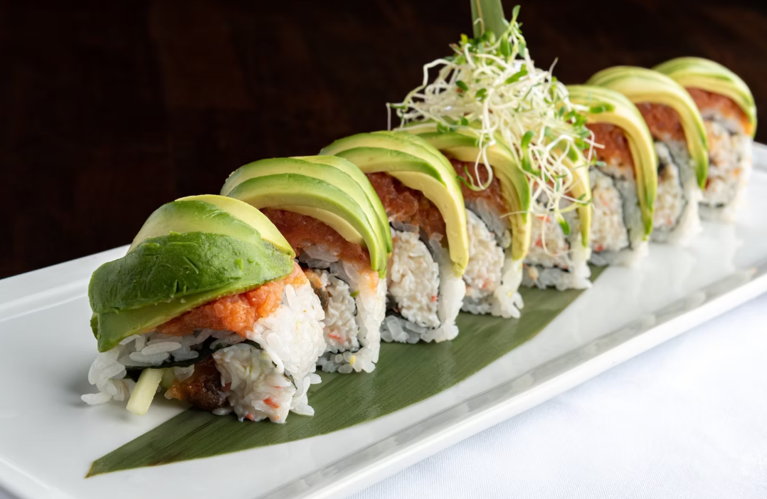 Sushi roll with avocado slices and shredded garnish on top, served on a white rectangular plate.