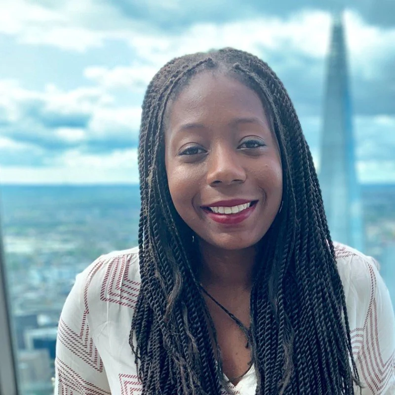 A woman with braided hair smiling in front of a cityscape and cloudy sky.