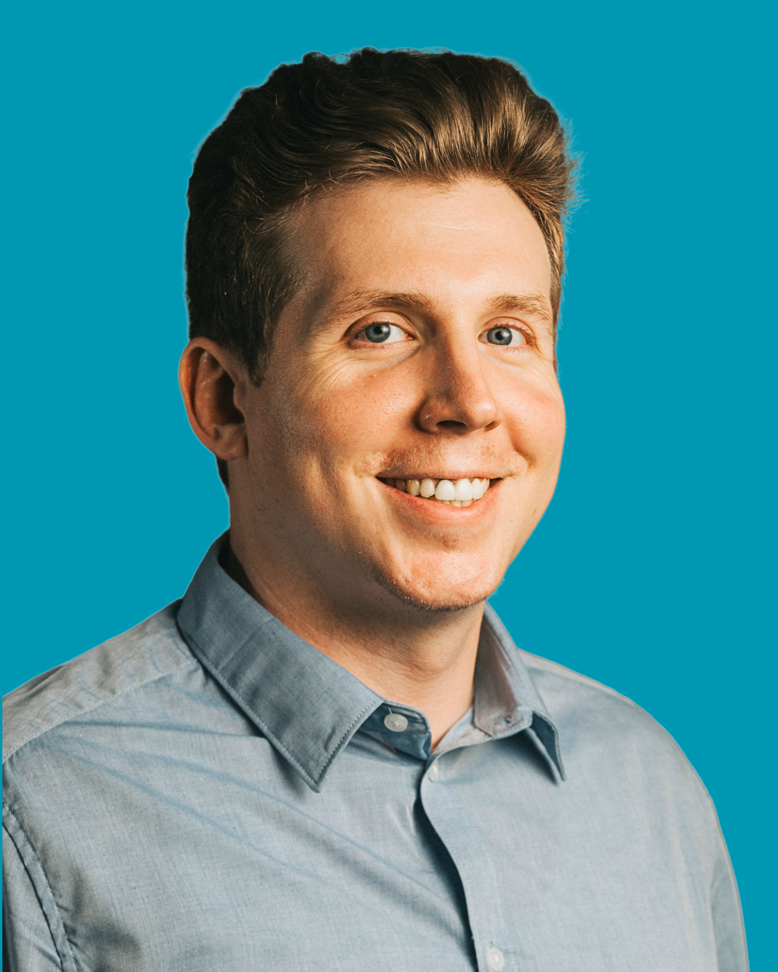 A young man with short brown hair, blue eyes, and light skin, smiling in front of a blue background, wearing a light blue collared shirt.