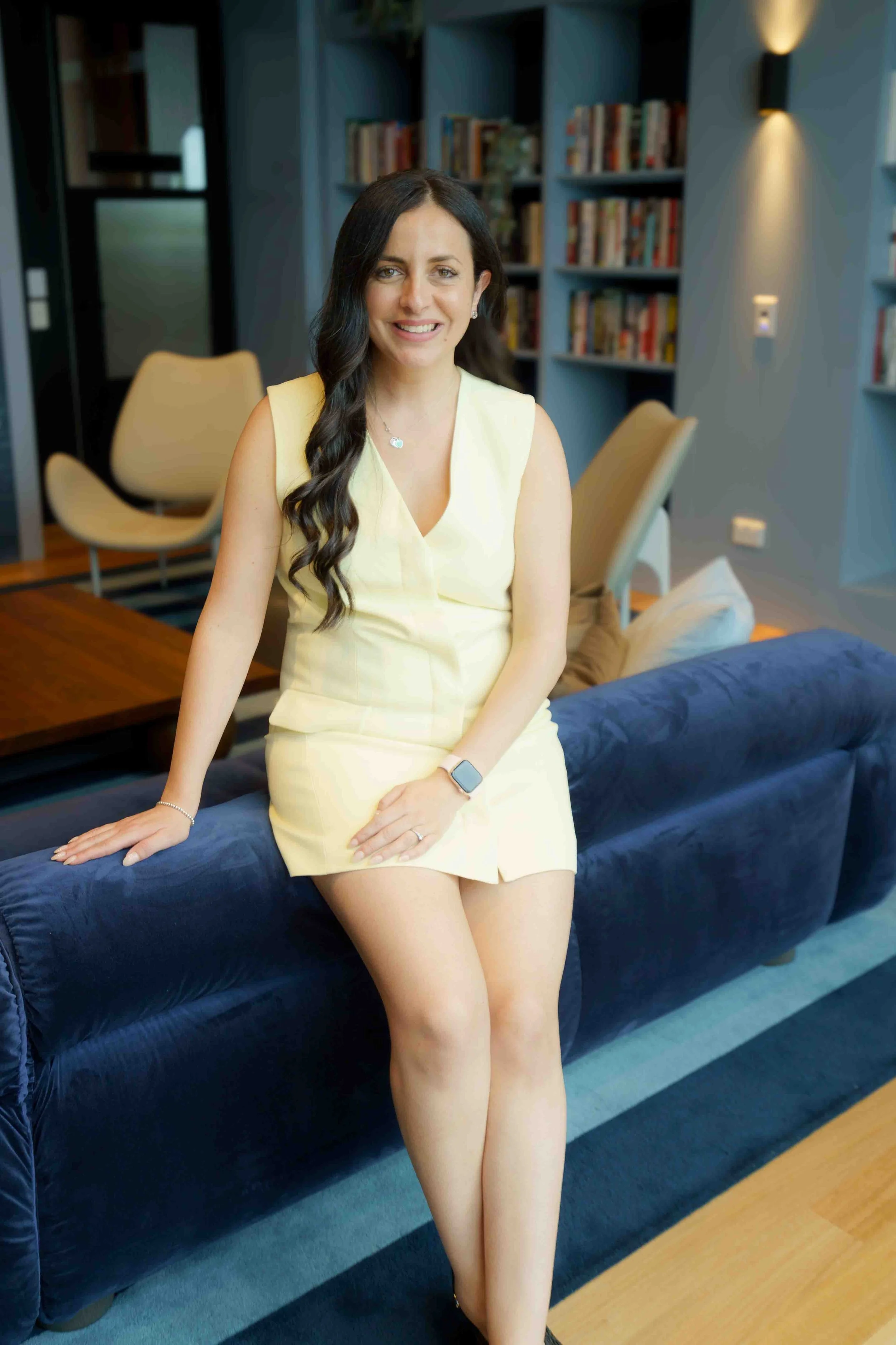 A woman with long dark hair, wearing a sleeveless yellow dress, sitting on a navy blue sofa in a modern living room with bookshelves in the background.