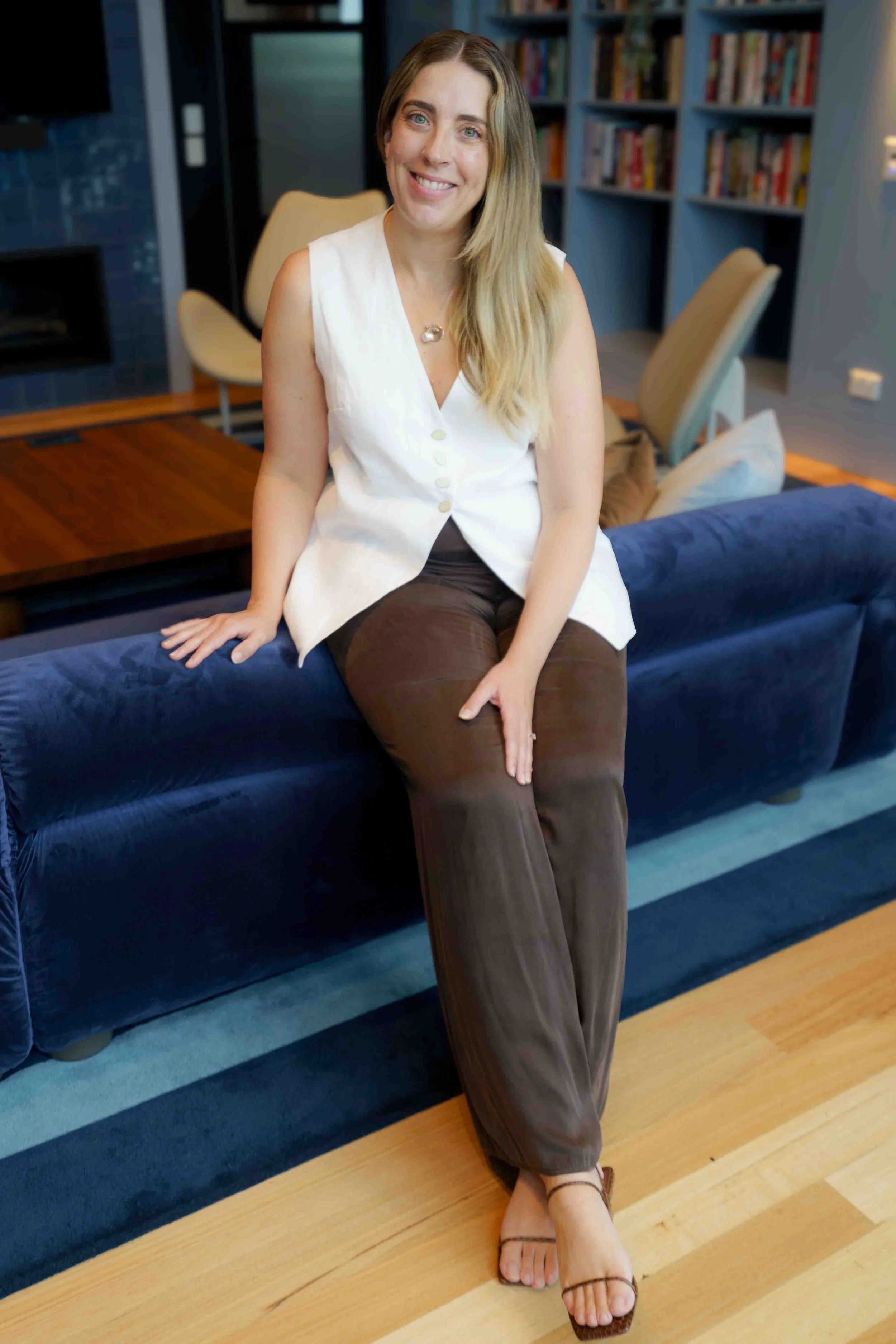 A woman with long blonde hair and a white sleeveless top sitting on a blue couch in a room with bookshelves and wooden furniture, smiling at the camera.