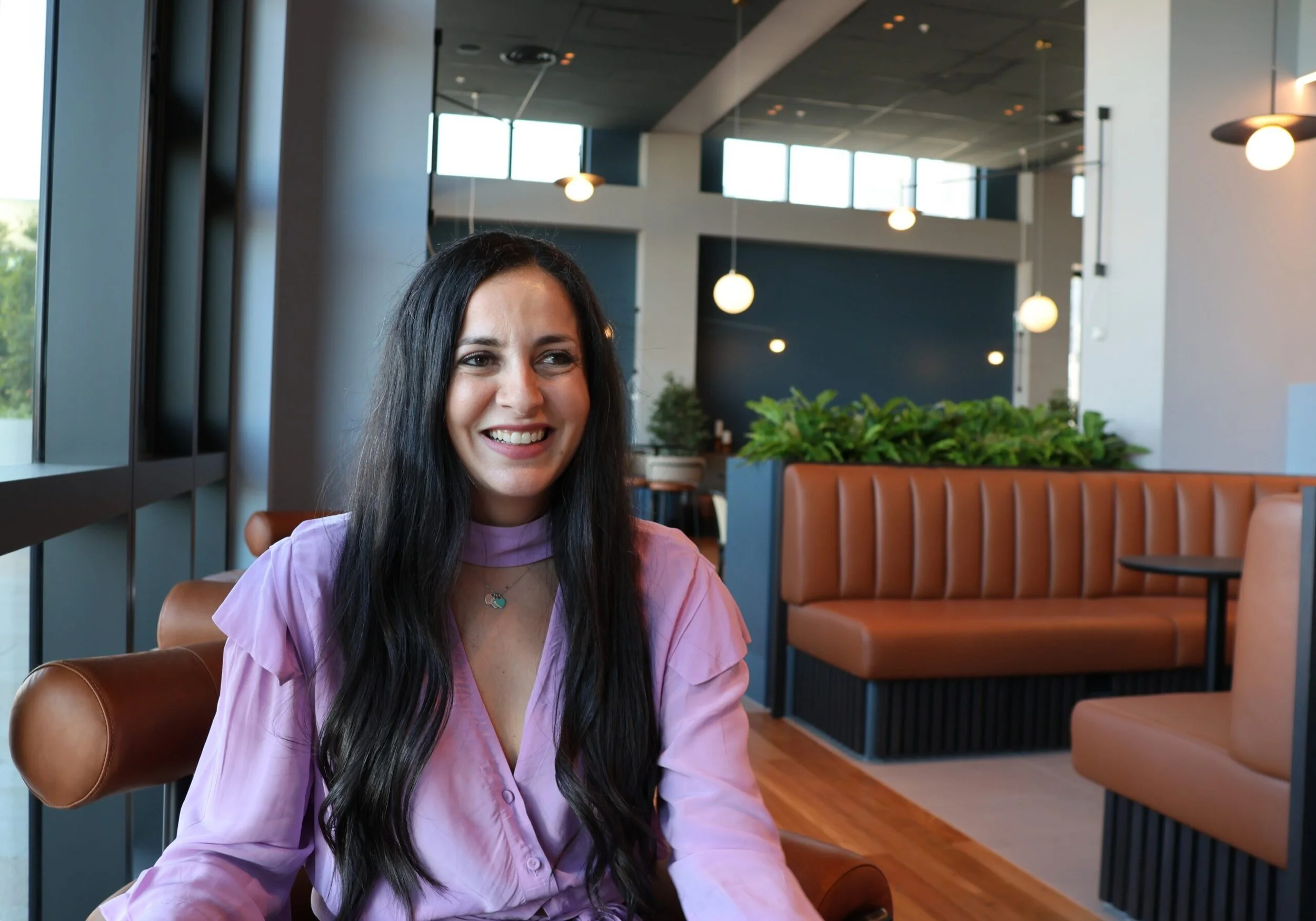 A smiling woman with long dark hair sitting inside a modern cafe with large windows, brown leather seats, and green plants in the background.