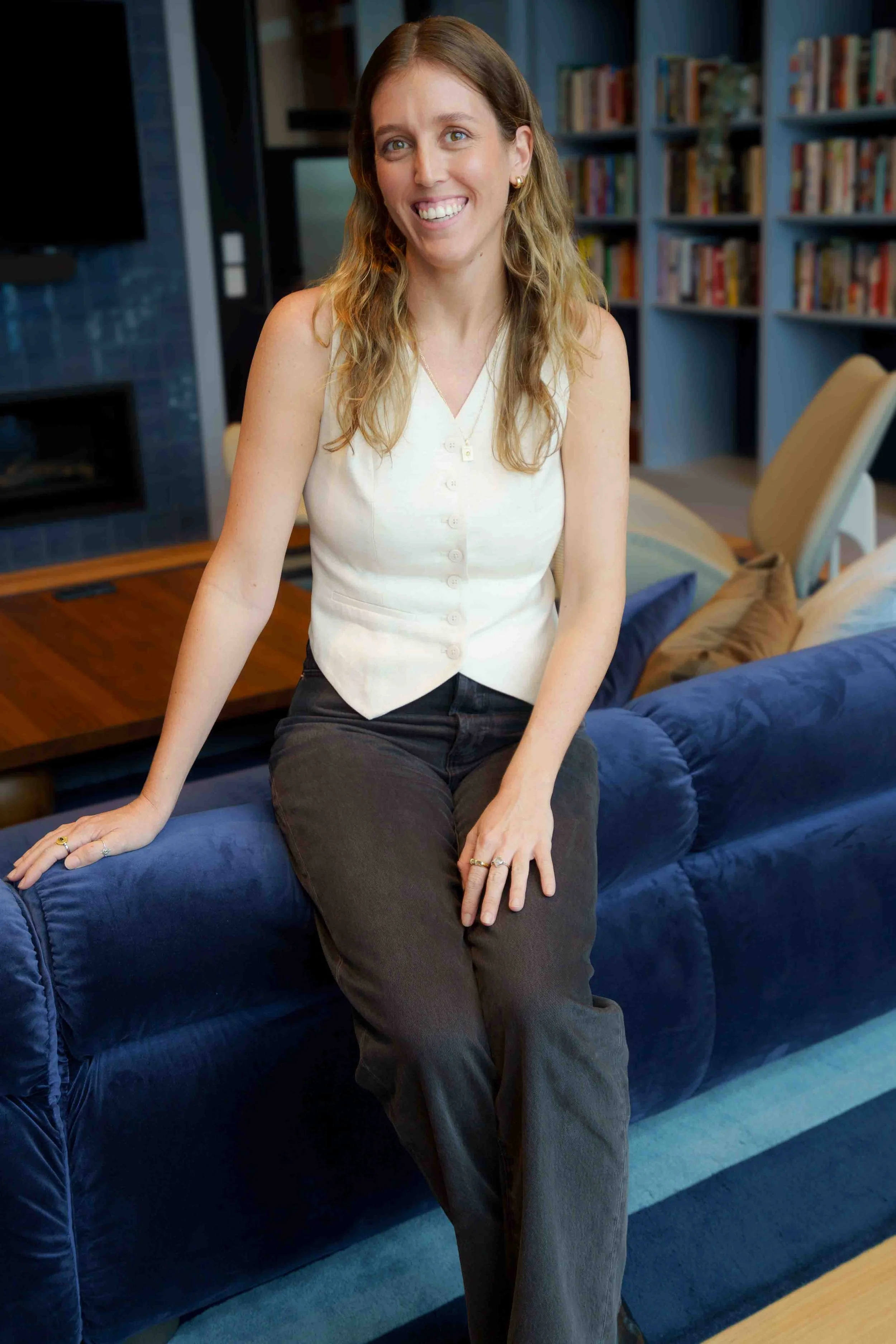 A woman with long wavy blonde hair, smiling, sitting on the arm of a blue sofa in a living room with bookshelves and a fireplace in the background.