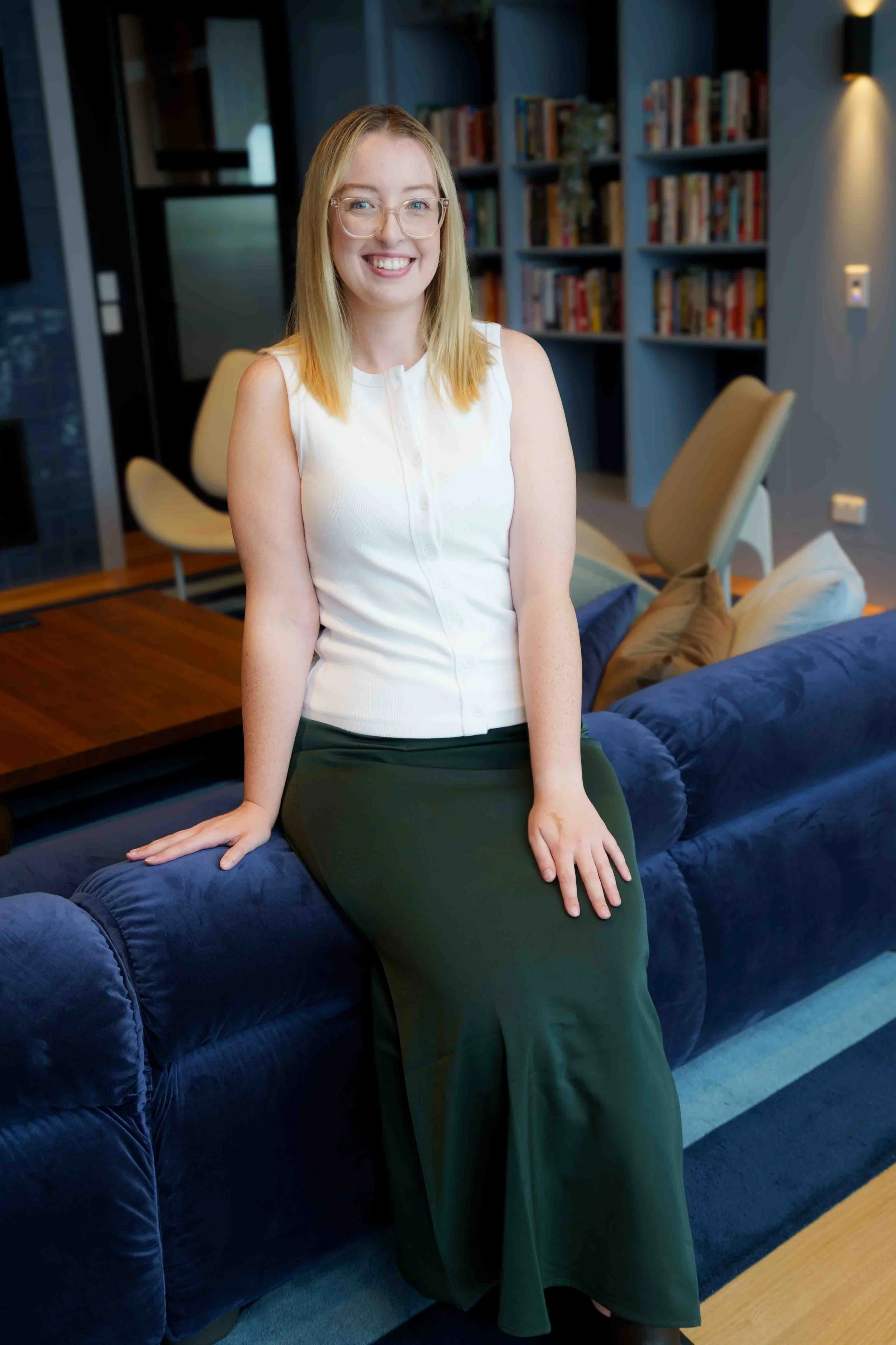 A woman with blonde hair, glasses, and a white sleeveless top sitting on a blue sofa in a library or living room, smiling at the camera.