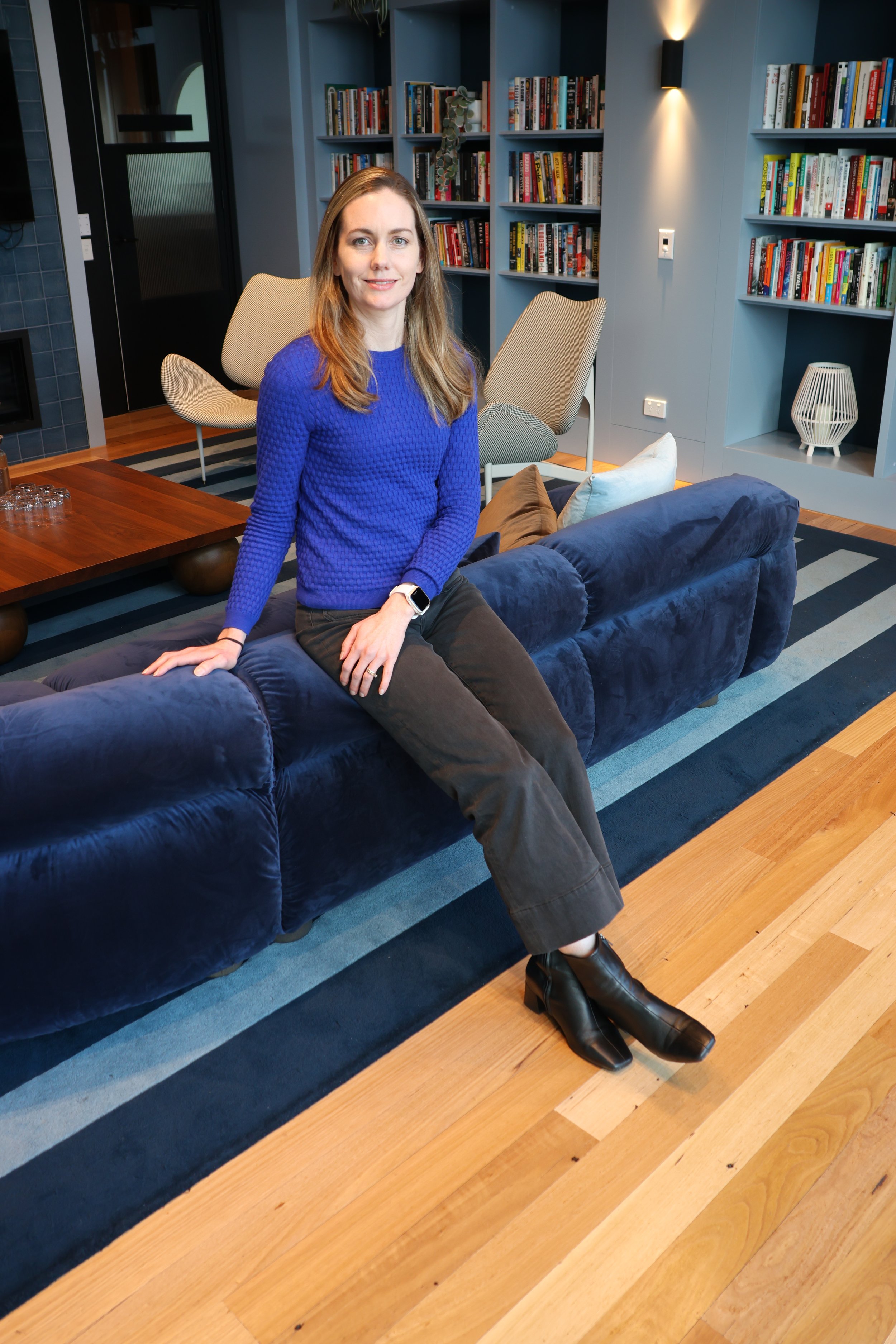 A woman sitting on a navy blue velvet sofa in a modern living room with bookshelves, a coffee table, and various pillows.