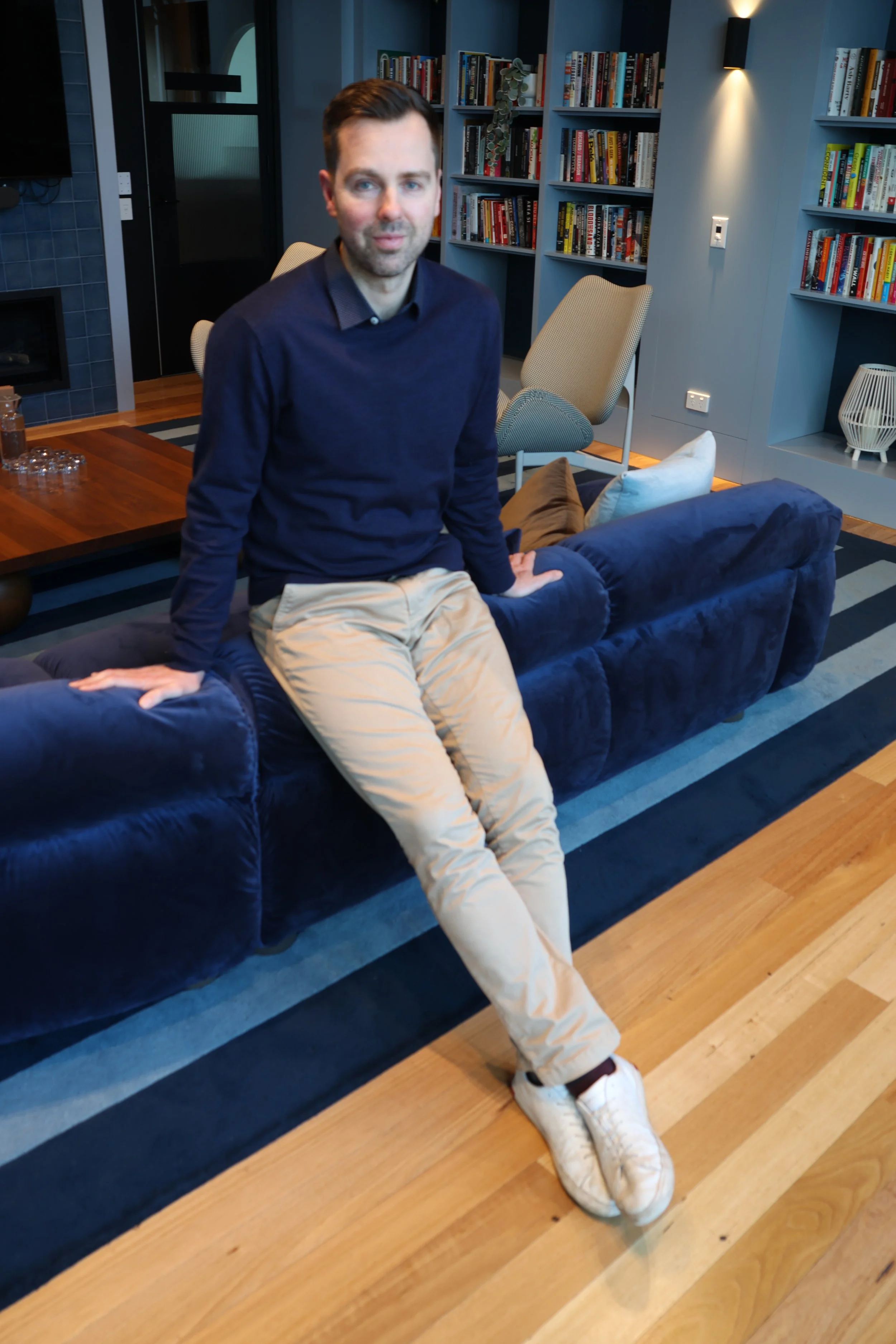 A man sitting on a navy blue sofa in a living room with bookshelves in the background.