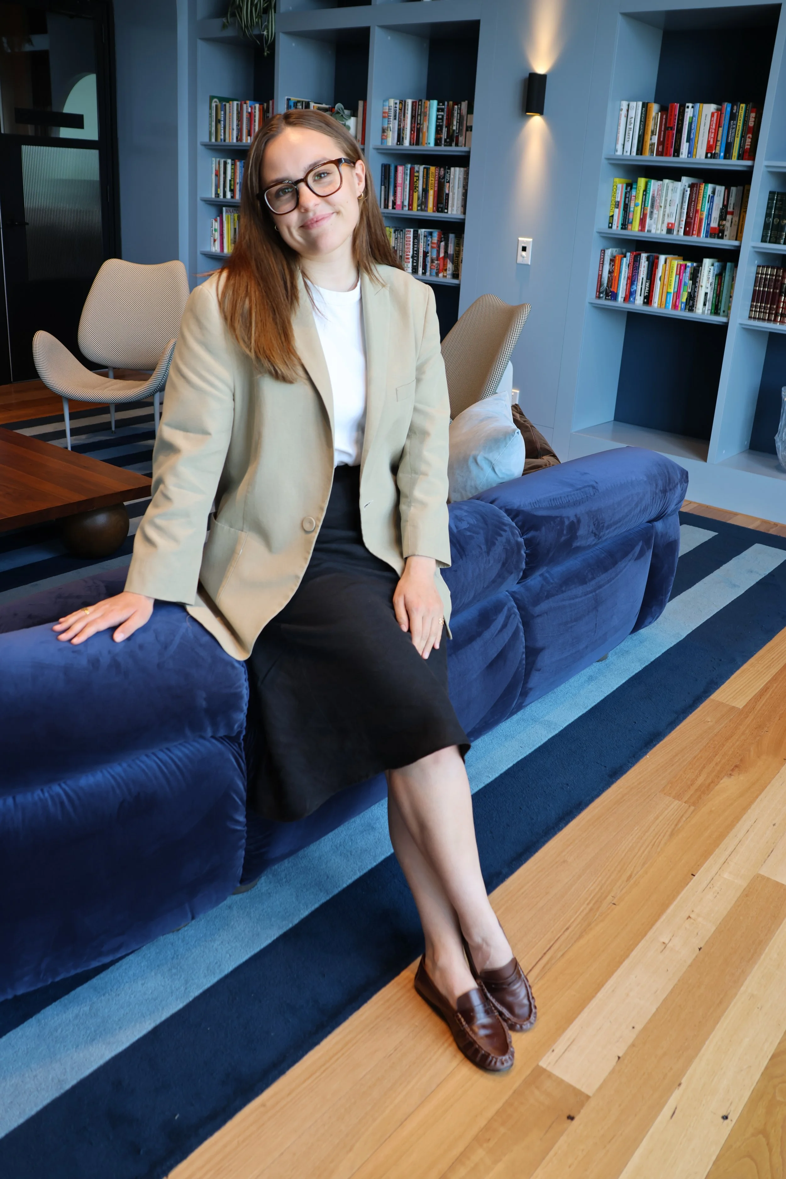 A woman with glasses sitting on a blue velvet couch in a modern library or lounge area with bookshelves behind her.
