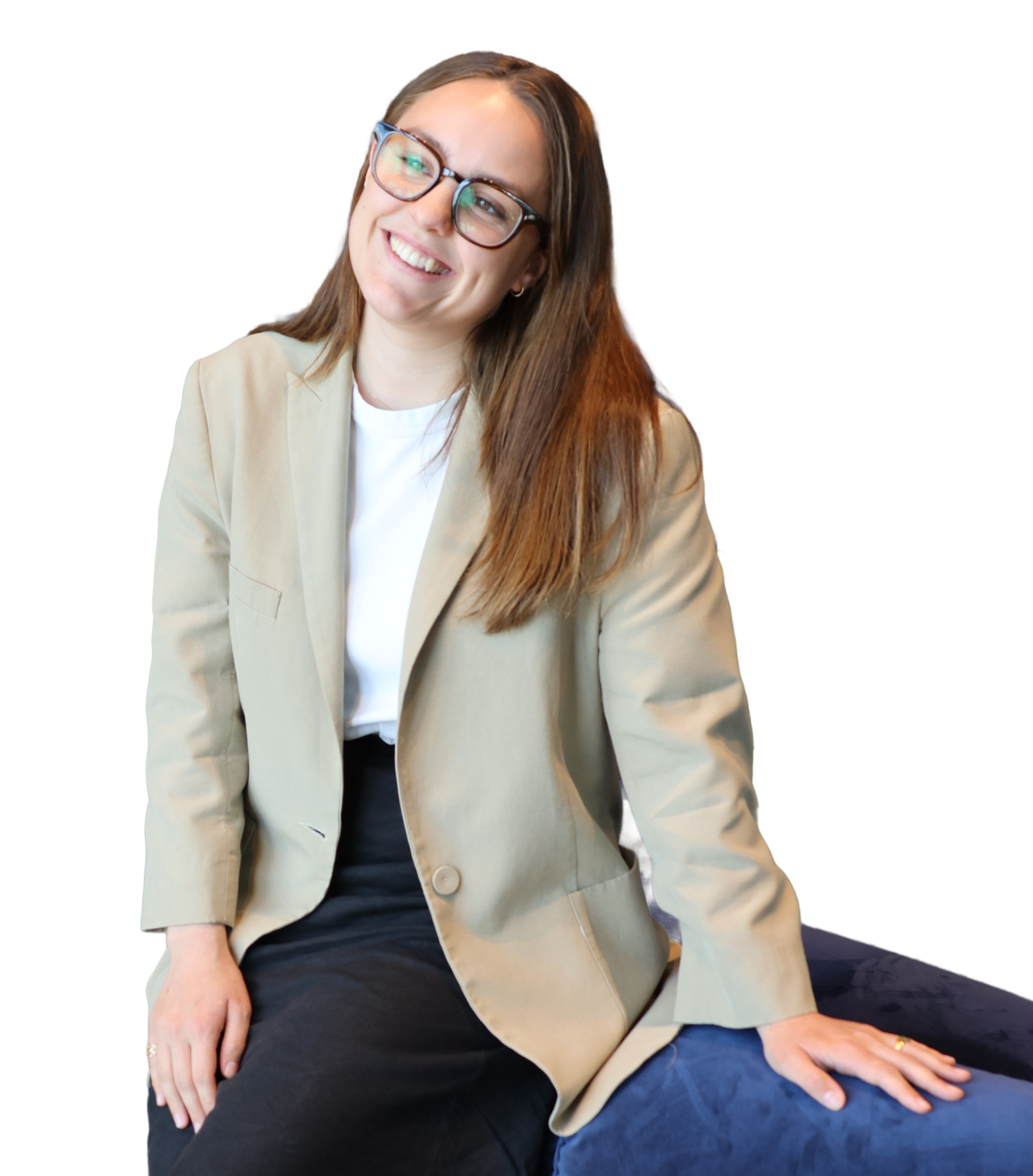 A woman wearing glasses and a beige blazer, sitting on a dark surface, smiling and looking slightly to the side.
