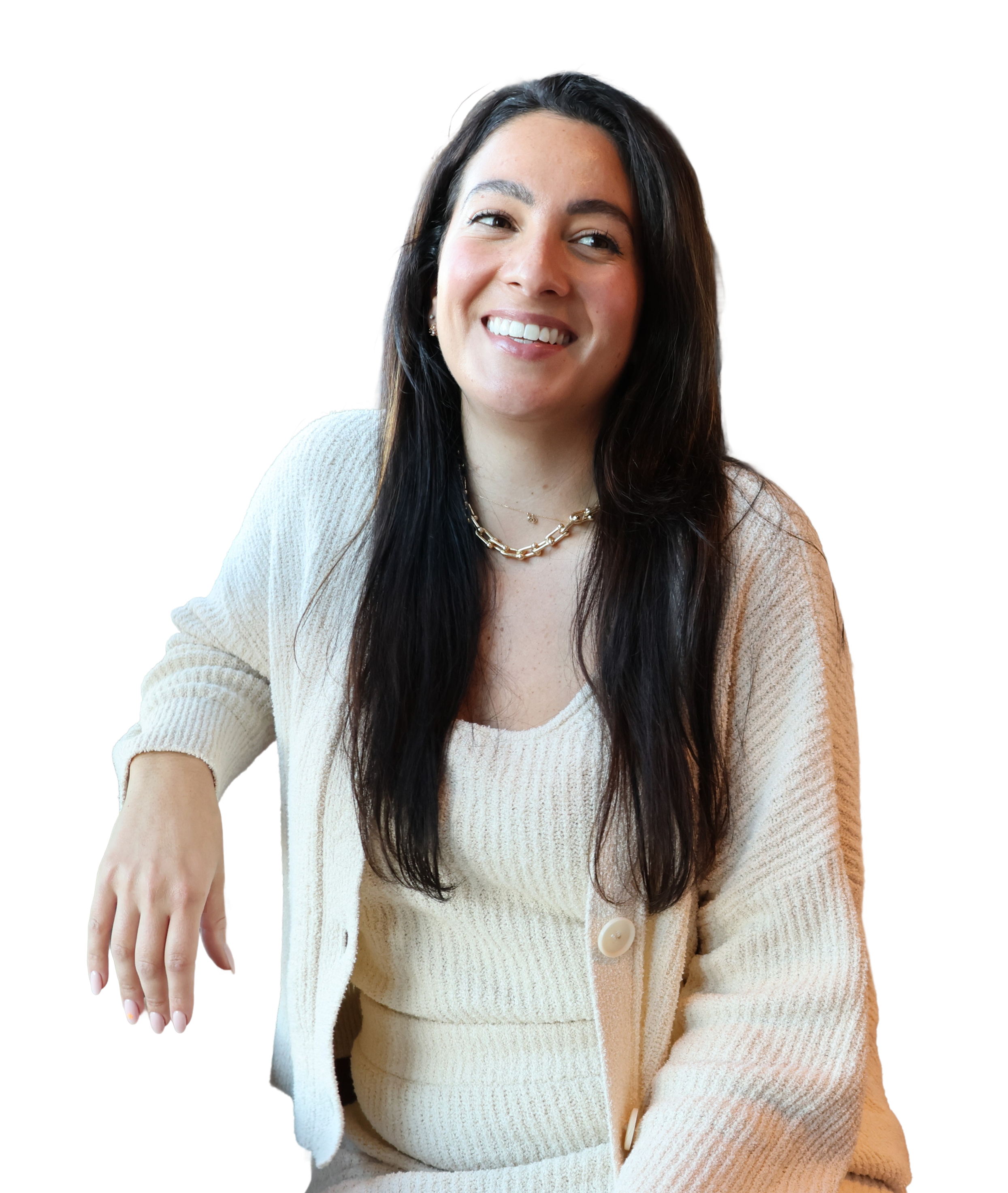 A woman with long dark hair wearing a light-colored cardigan and a gold chain necklace, smiling and sitting with her hand resting on a surface.