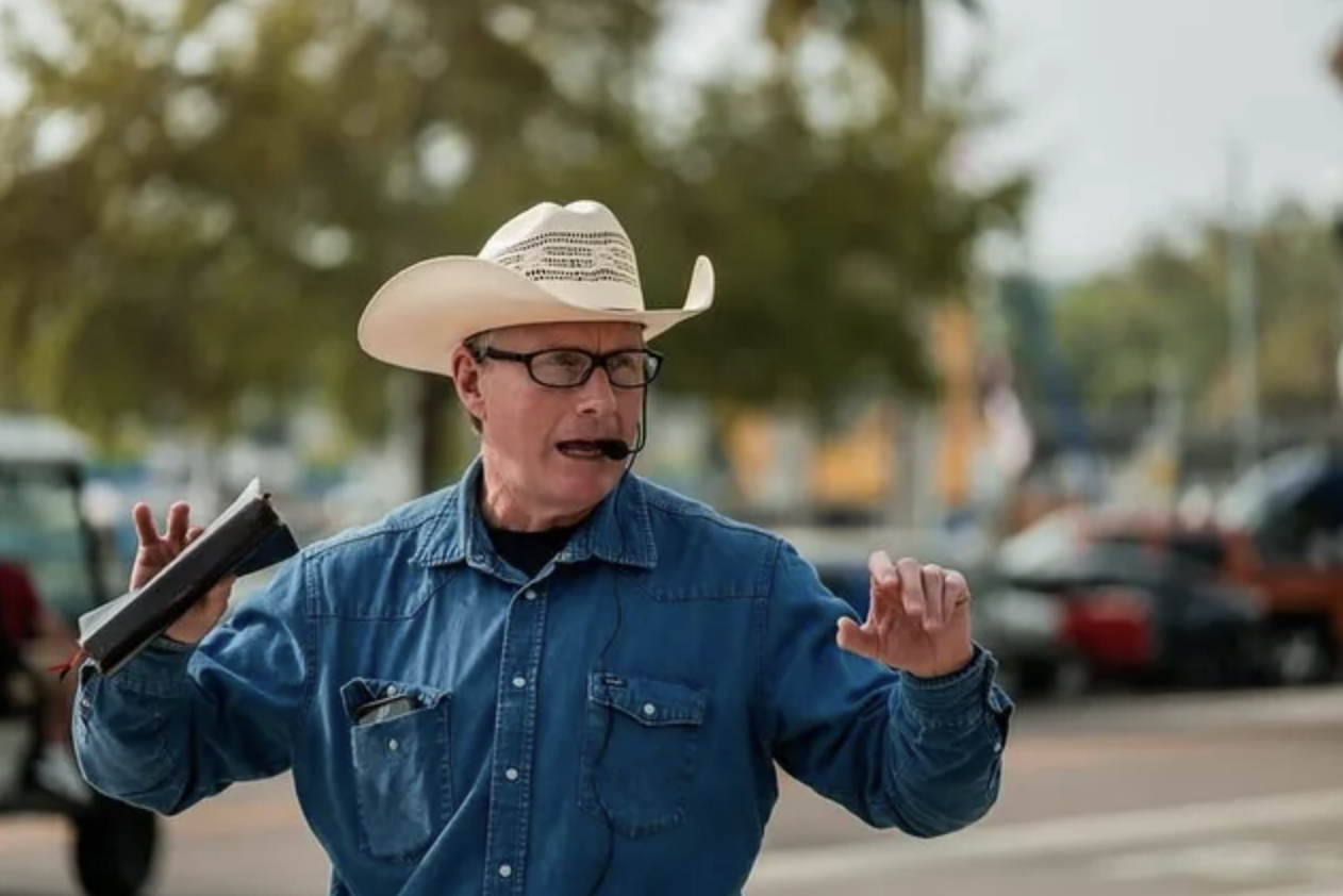 A man wearing a cowboy hat, glasses, and a denim shirt, speaking passionately with a headset microphone, gesturing with one hand, outdoors with parked cars and trees in the background.