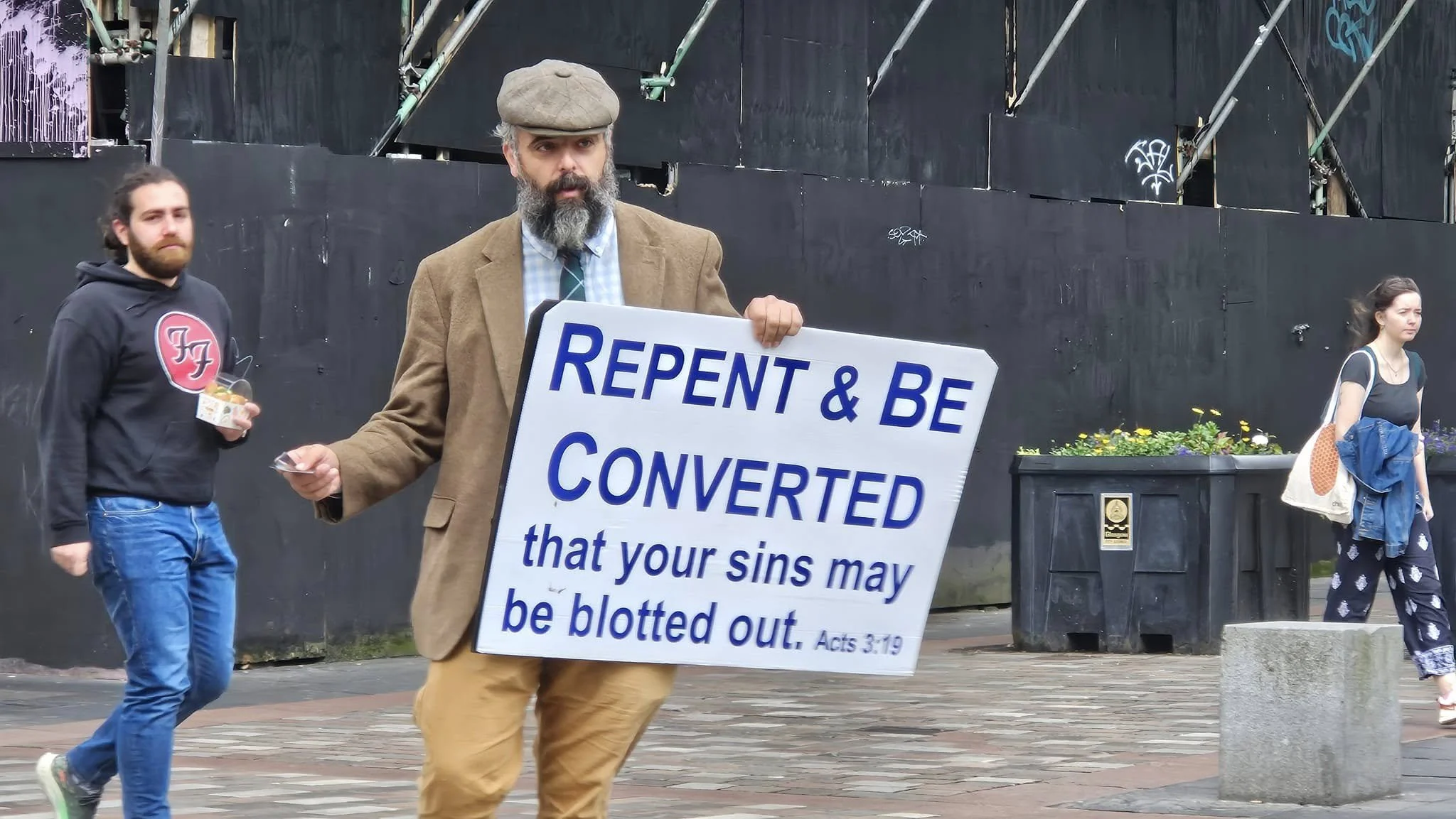 A man holding a sign that reads 'RENP & BE CONVERTED that your sins may be blotted out. Acts 3:19' walks along a street. Two other people are visible in the background, a man in a black hoodie and blue pants holding a snack and a woman in patterned pants carrying a bag.
