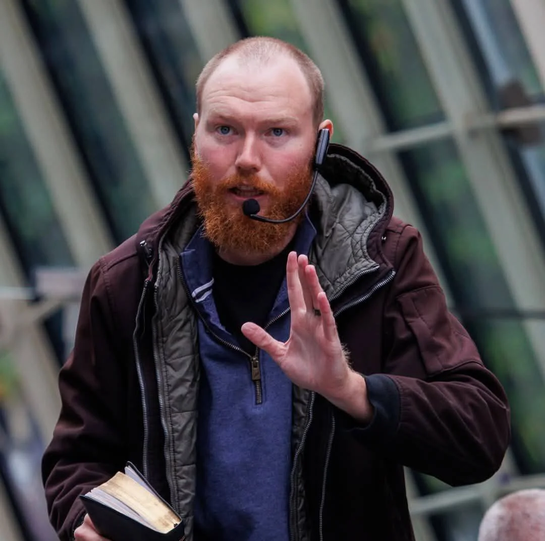 A man with a red beard and light skin wearing a headset microphone, holding a book, and gesturing with his hand.
