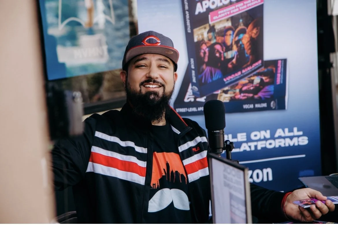 A man with a beard and a cap smiling at a camera, standing in front of a promotional poster for a show called 'APOLLO' and holding a phone in his hand.