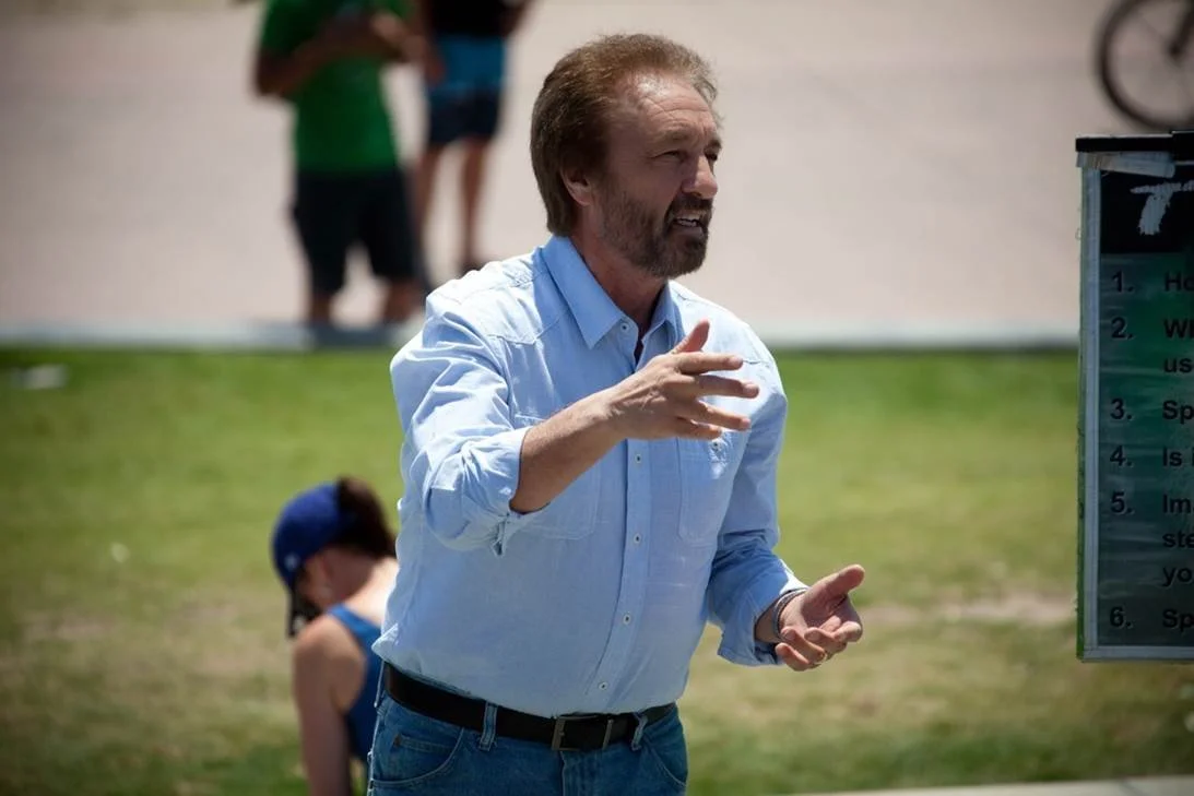 A man with light brown hair and a beard, wearing a light blue button-up shirt and jeans, is speaking outdoors in front of a grassy area and a signboard. In the background, there are children and blurred figures, indicating a public or community setting.
