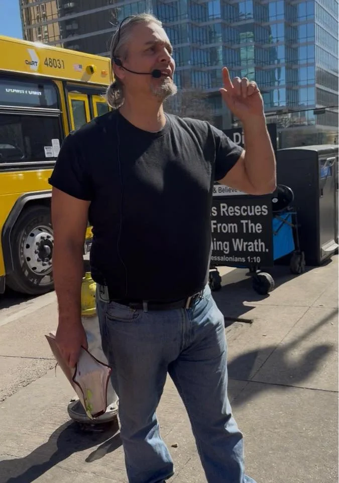 A man with gray hair, beard, wearing a black t-shirt and jeans, speaking into a microphone headset while holding papers, standing outdoors in front of a yellow bus and a sign about rescuing from God's wrath.