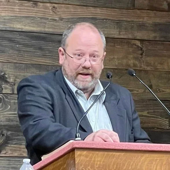 Man with glasses and beard speaking at a lectern in front of a wooden wall.