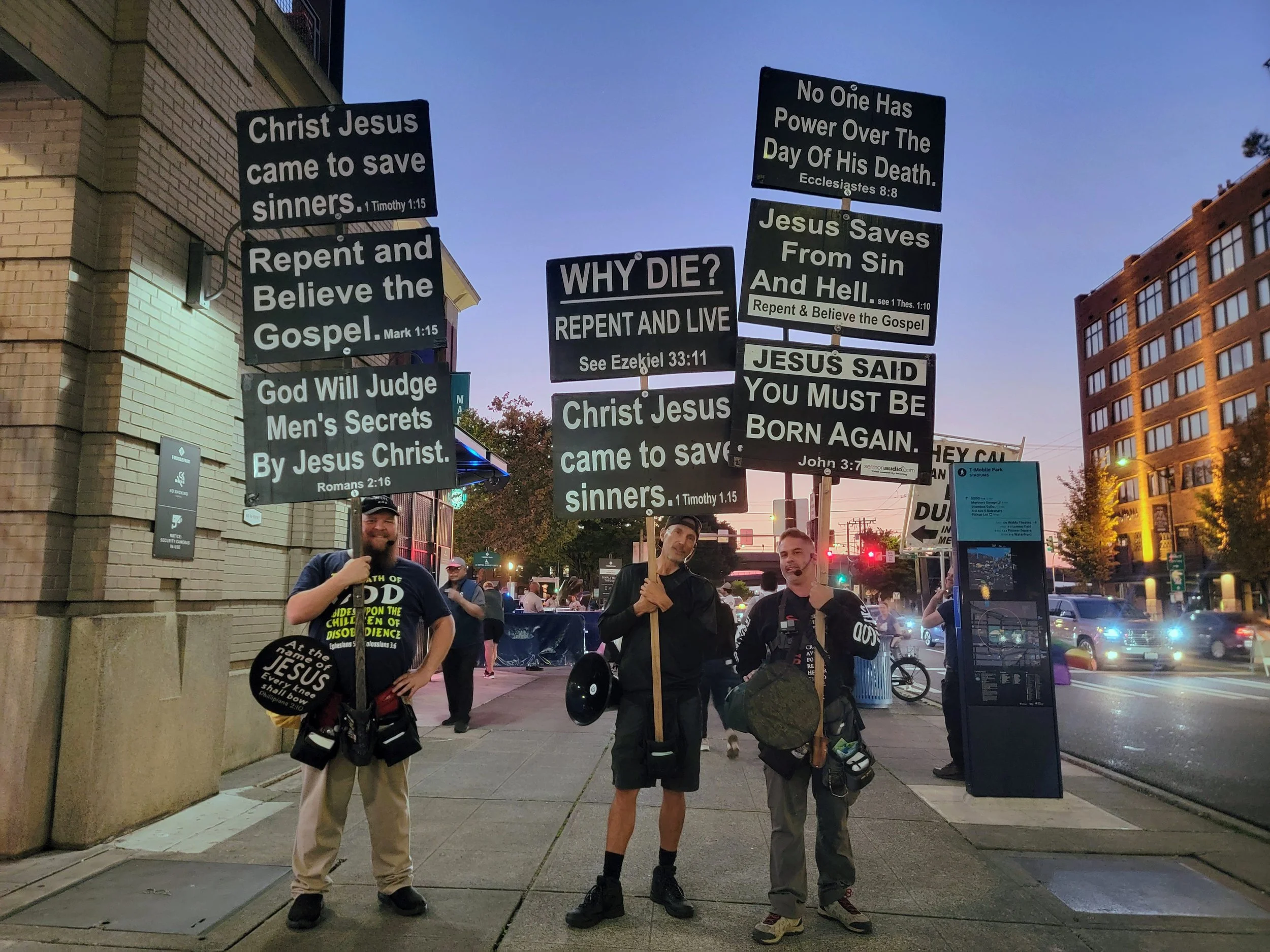 Three men standing on a city sidewalk holding protest signs with Christian messages, buildings and traffic in the background during dusk.