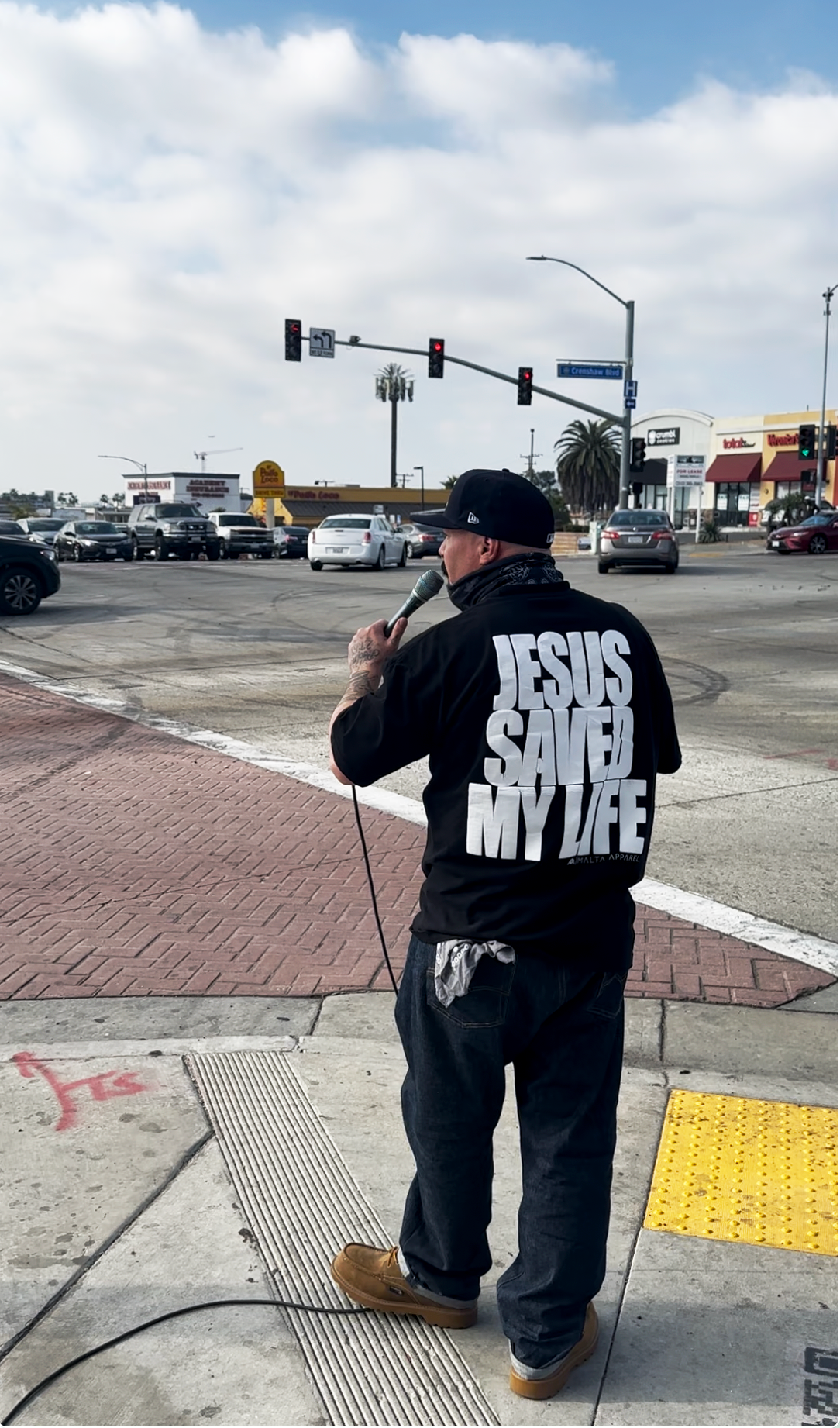 A man wearing a black hoodie with the message 'Jesus Saved My Life' on the back, holding a microphone and speaking on the sidewalk at an urban crosswalk, with cars and stores in the background.