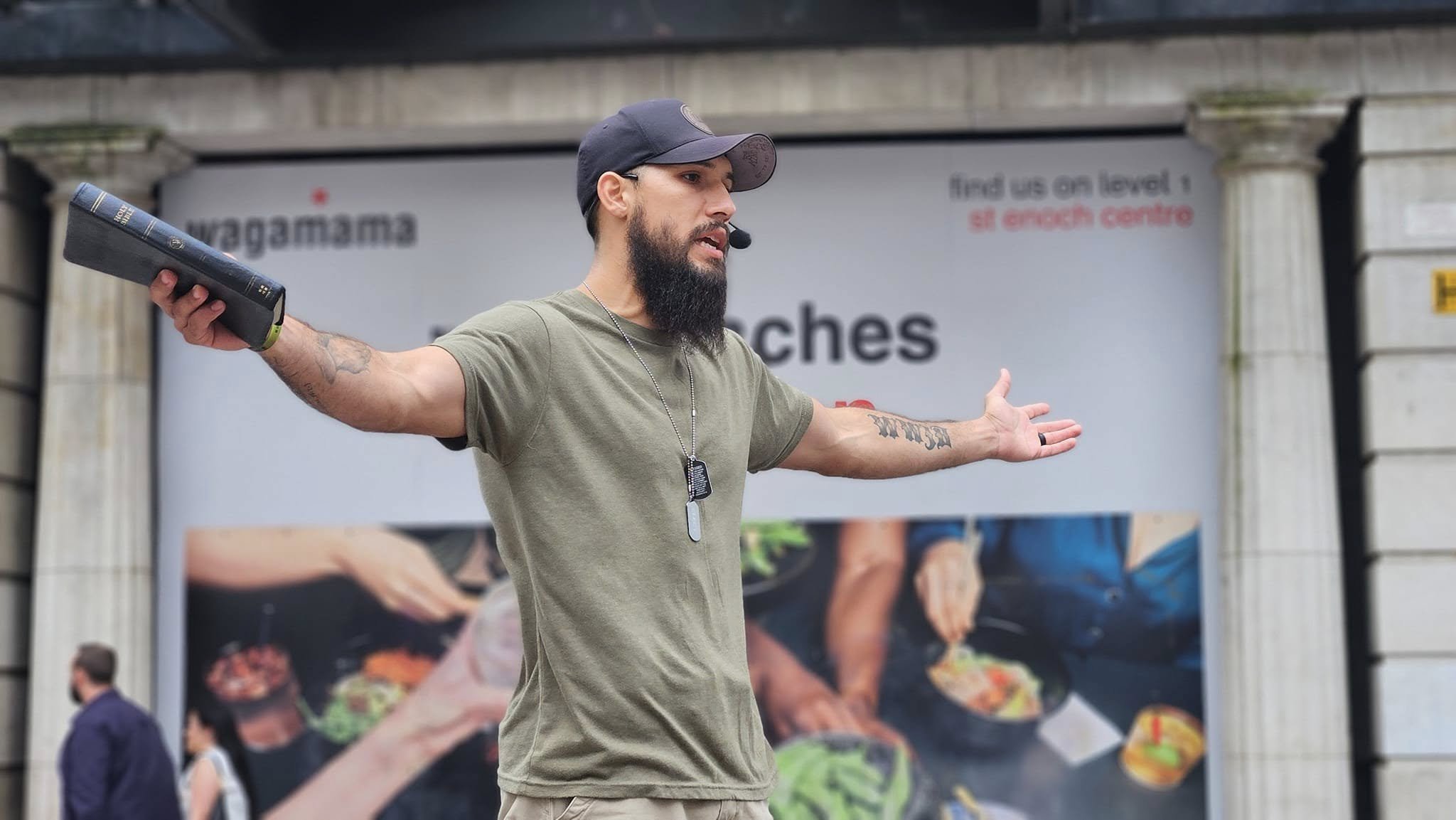 Man with beard, wearing a black cap, microphone headset, and dog tags, gestures with arms outstretched on a busy sidewalk in front of a large billboard.