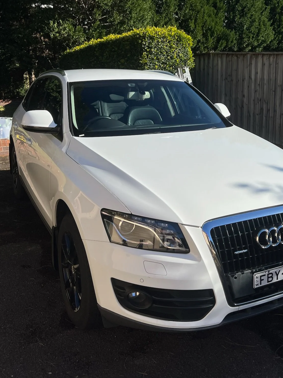 White Audi sedan parked outdoors, showing the front and driver's side, with a tree and wooden fence in the background.