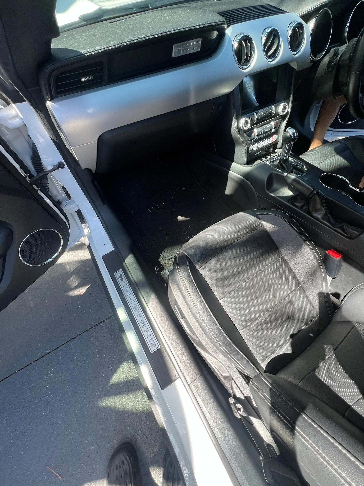 Inside view of a Ford Mustang car with black seats, dashboard, and center console, with the driver's side door open revealing treadmarks and a person’s foot in crocs.