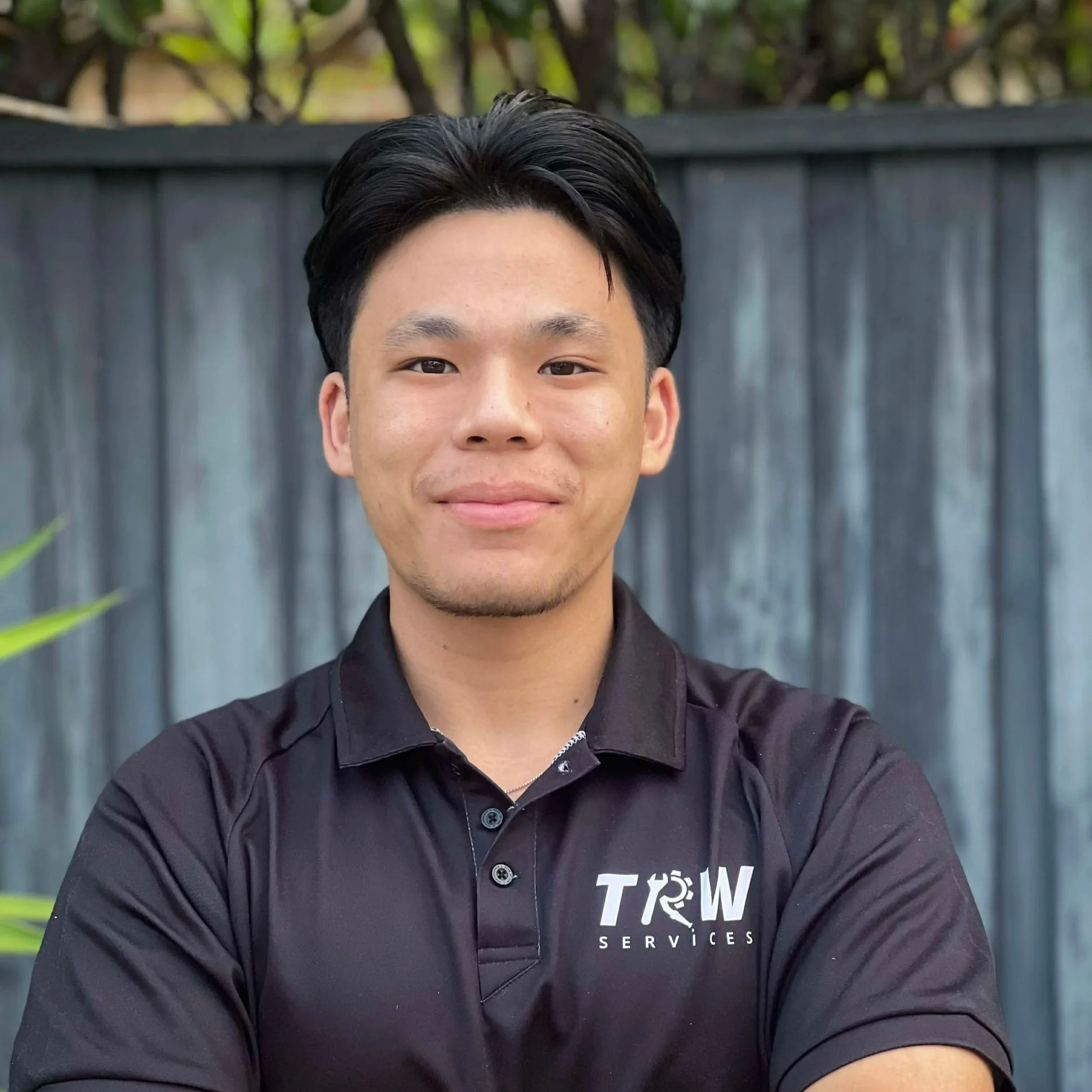 Portrait of a young Asian man with black hair, wearing a black polo shirt with 'TOW SERVICES' logo, standing outdoors with a gray wooden fence in the background.