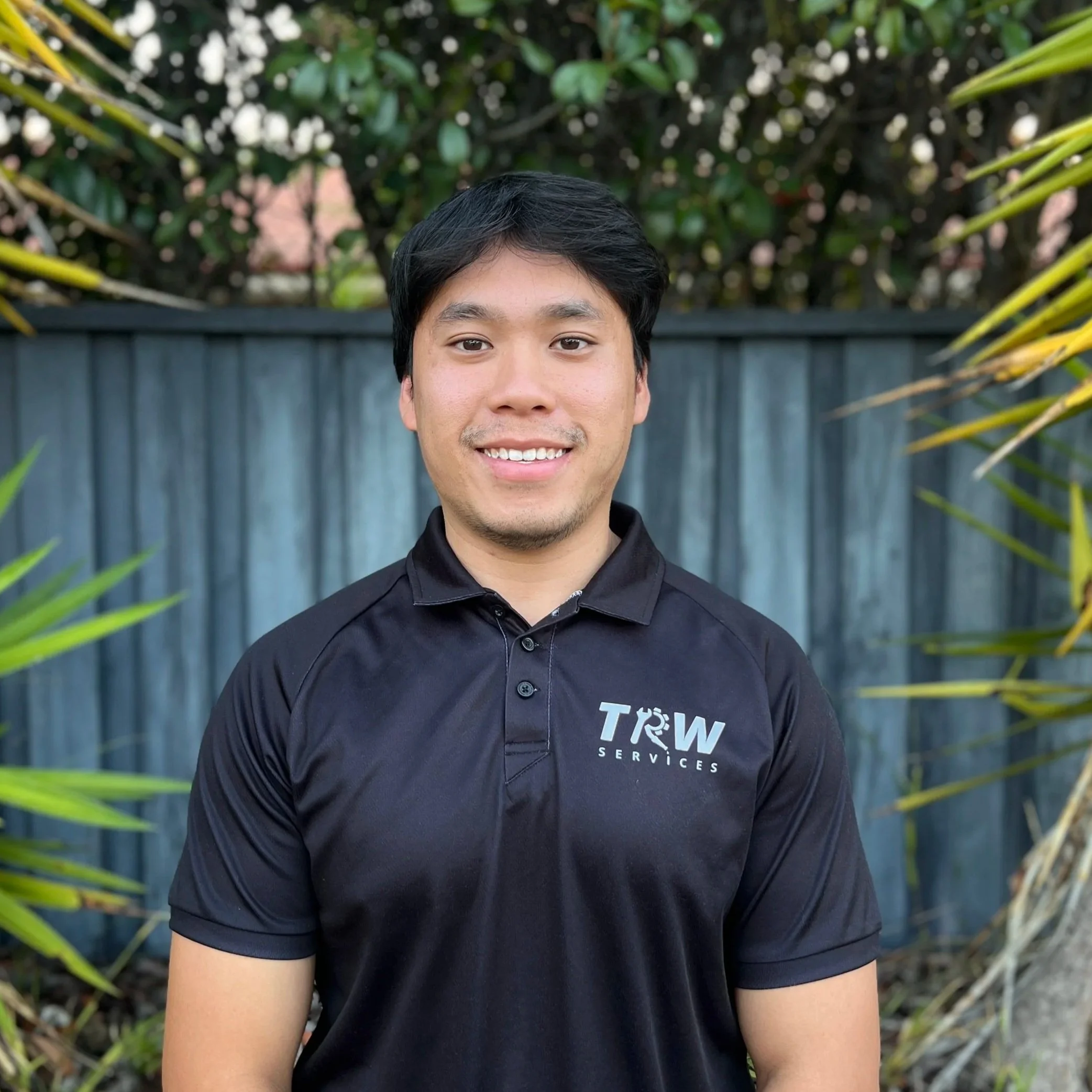A smiling young man with dark hair wearing a black polo shirt with the logo 'TRW Services' stands outdoors in front of a blue wooden fence, surrounded by green plants.