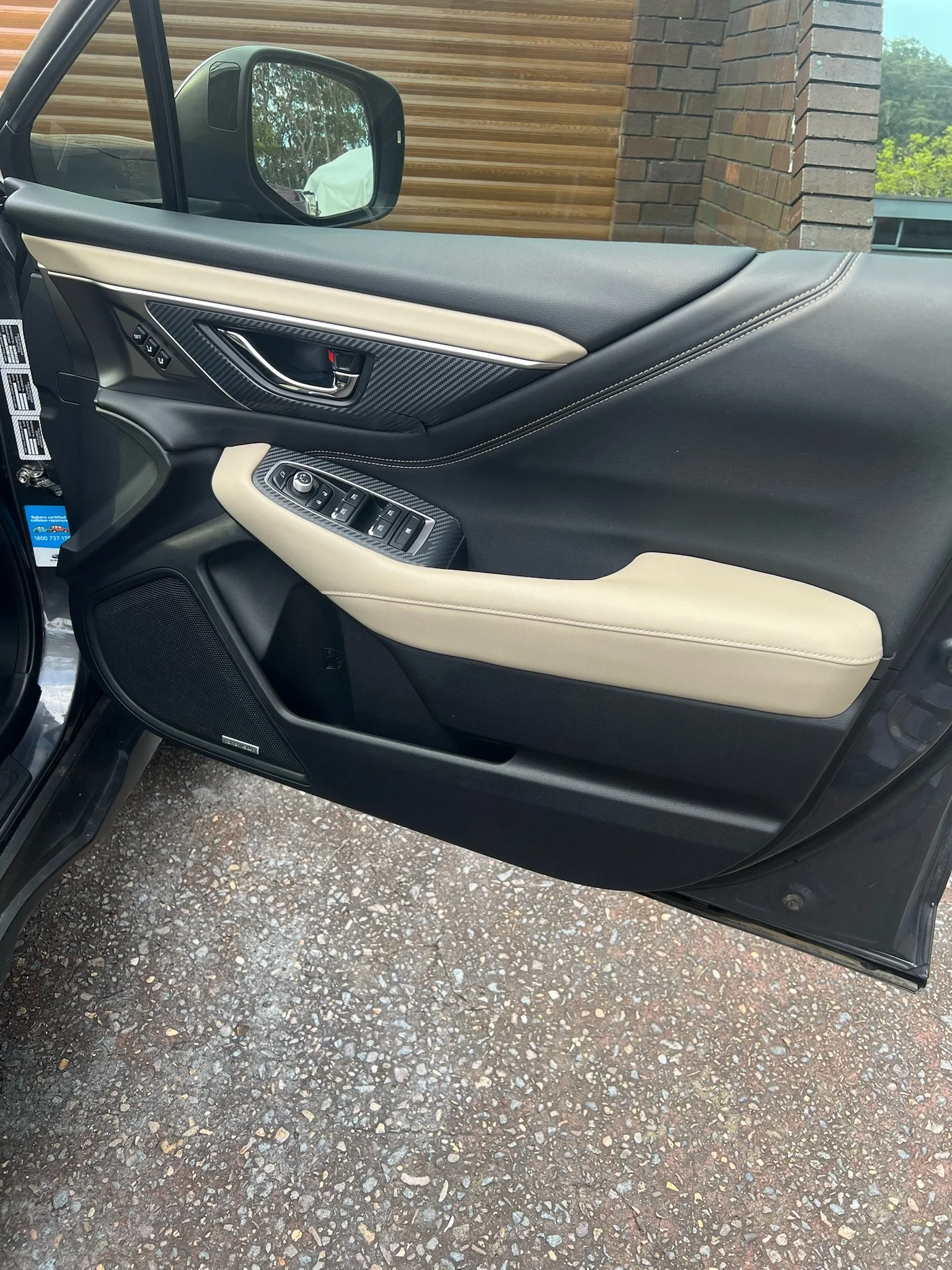 The interior of a car door featuring beige and black design with control buttons for windows and mirrors, a carbon fiber-patterned trim, a speaker, and a door handle, next to a side mirror and a brick wall outside.