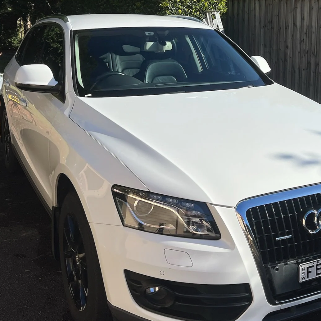 A white Audi car parked outdoors near a wooden fence with trees in the background.