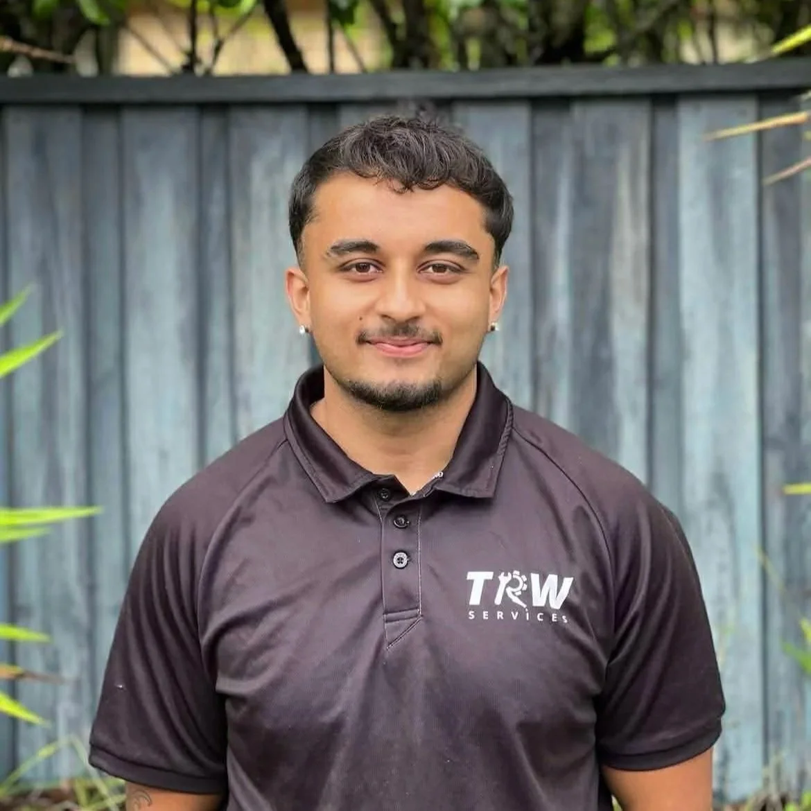 Young man with short dark hair, earrings, and a slight beard, wearing a black polo shirt with a company logo, standing outdoors in front of a wooden fence with plants nearby.