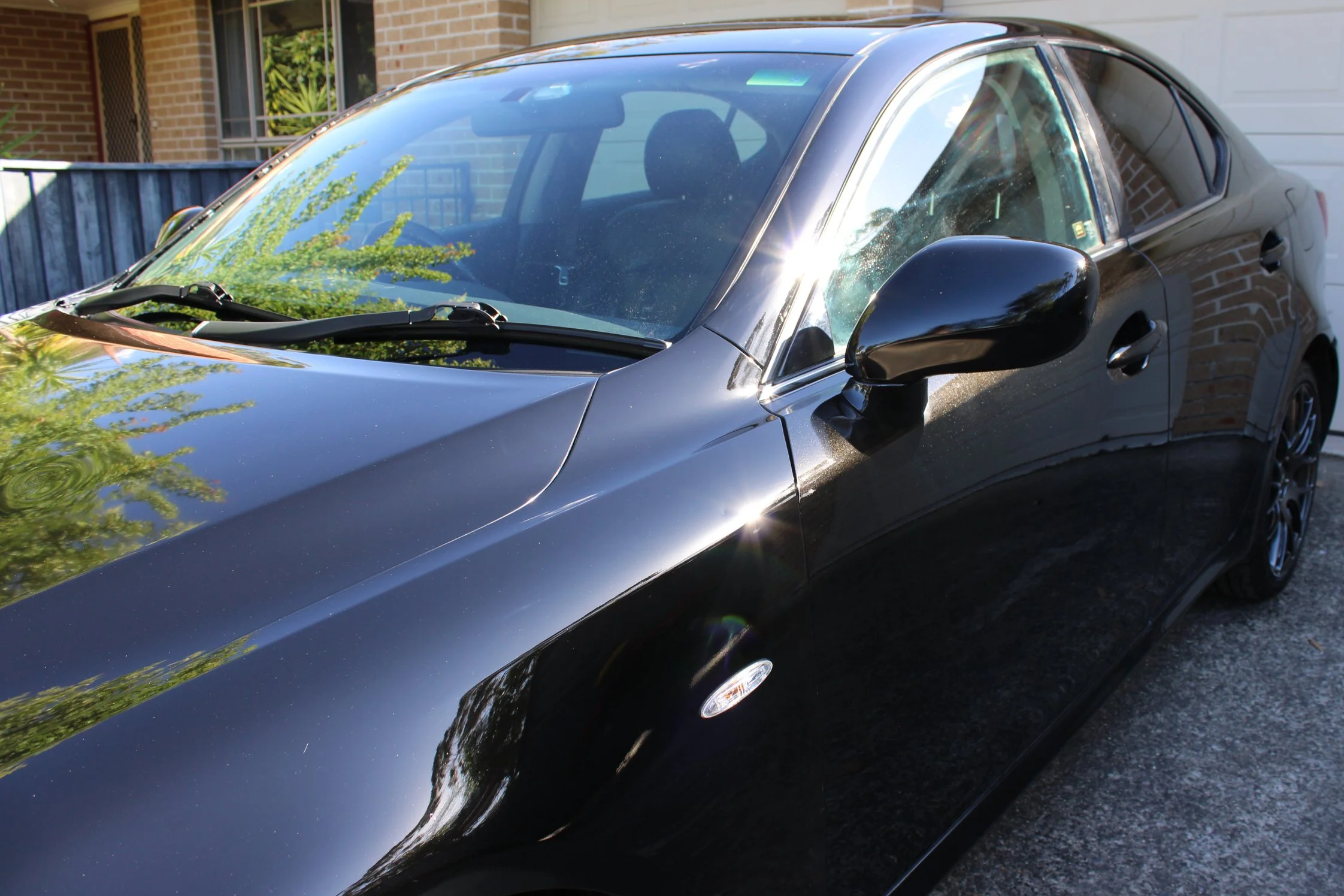 A black sports car parked in front of a house with a brick wall and a white garage door, with reflections of trees and the sky on its shiny surface.