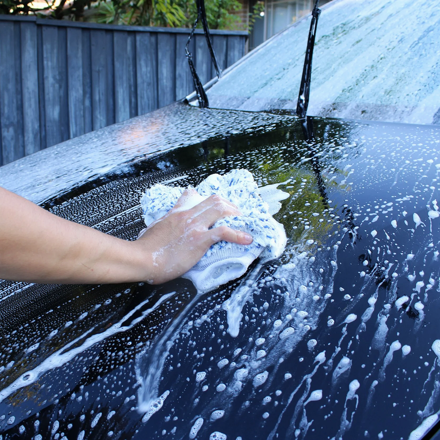 Person hand scrubbing the black car with a white sponge, washing away soap and suds in an outdoor driveway.