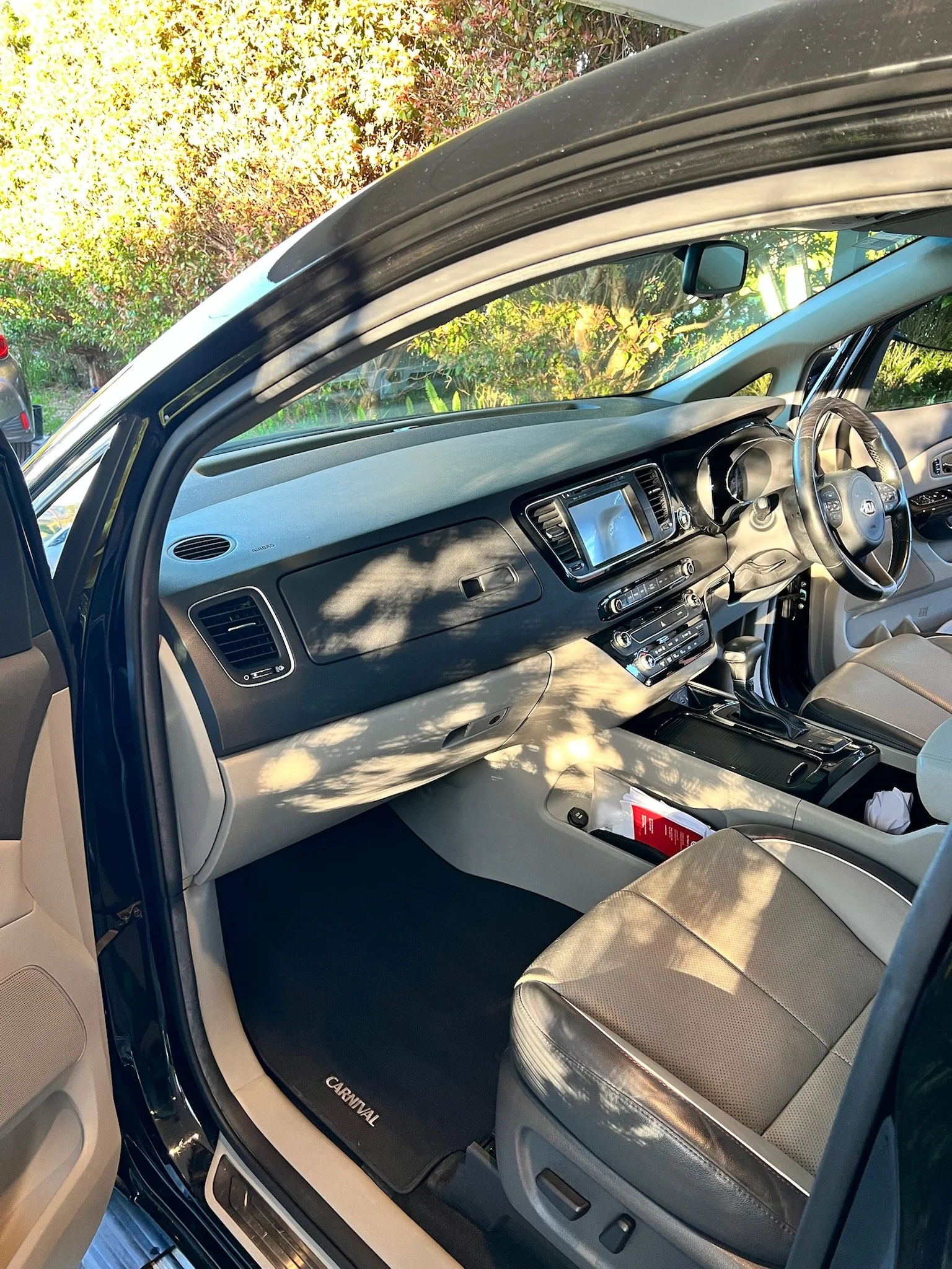 The interior of a Kia Carnival minivan showing the dashboard, steering wheel, center console, and front passenger seat, with sunlight and trees casting shadows inside.