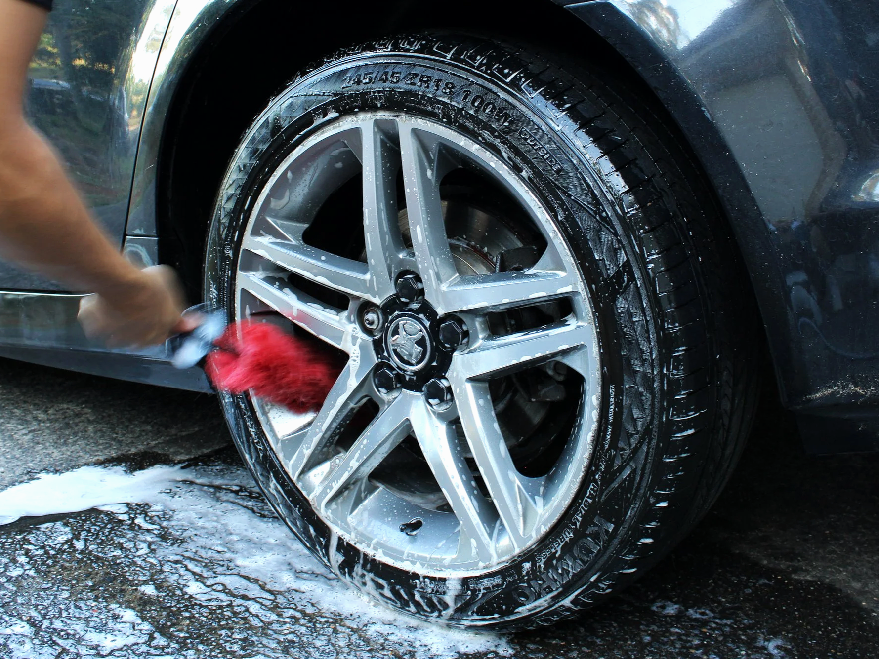 Person washing a black car's tire with a red sponge, soap suds and water are visible around the wheel.