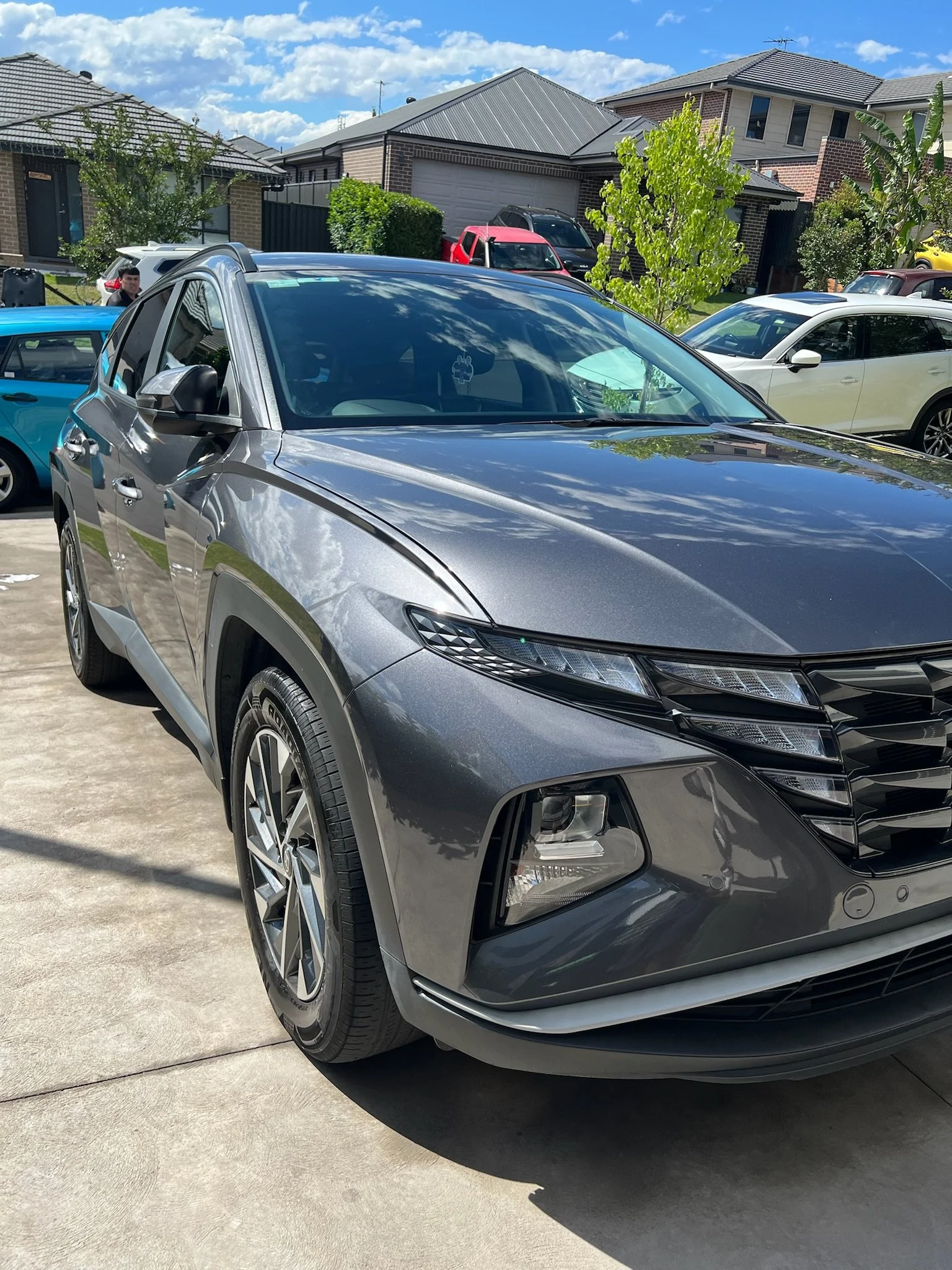 Front view of a metallic gray Honda SUV parked in a driveway with other cars and houses in the background.