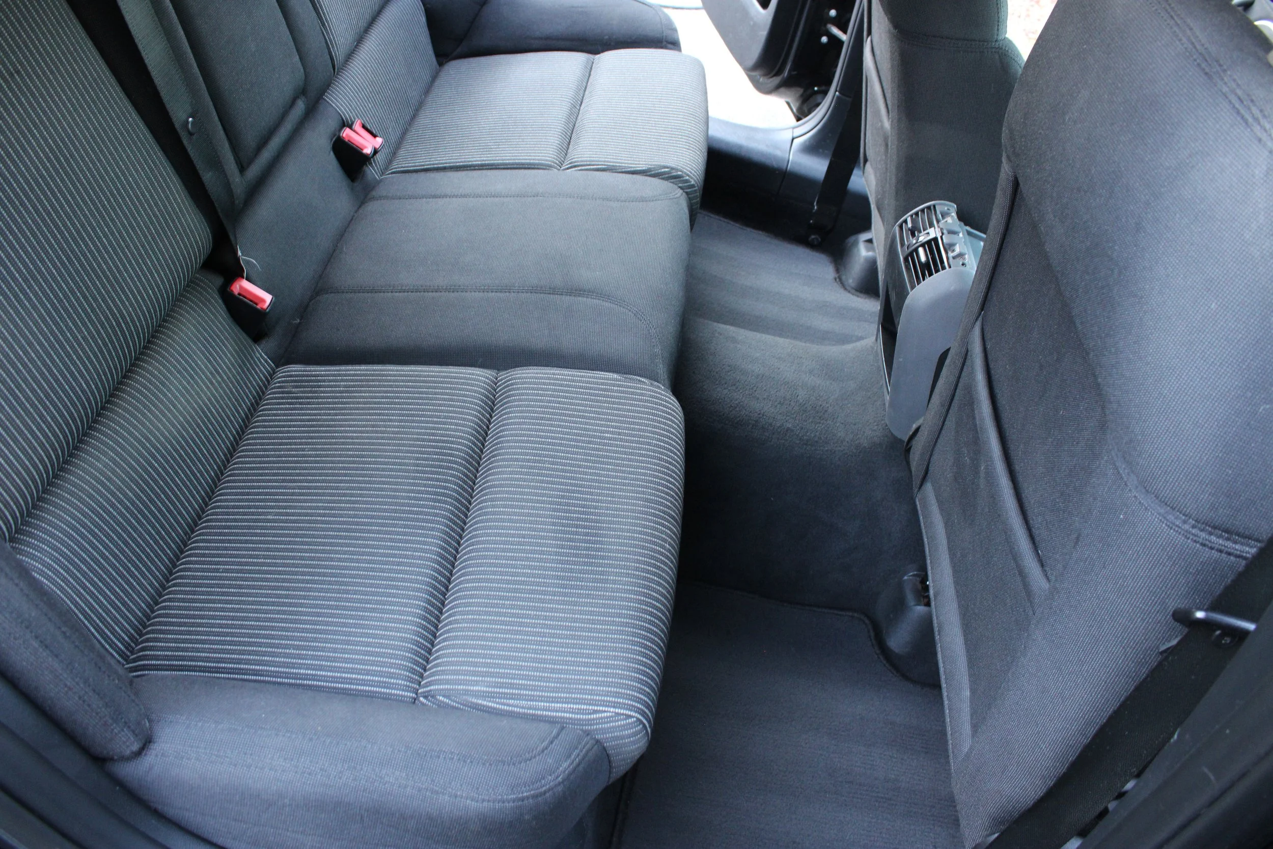 The backseat interior of a car with dark fabric seats, seat belts, and a rear air vent, viewed from the open rear door.