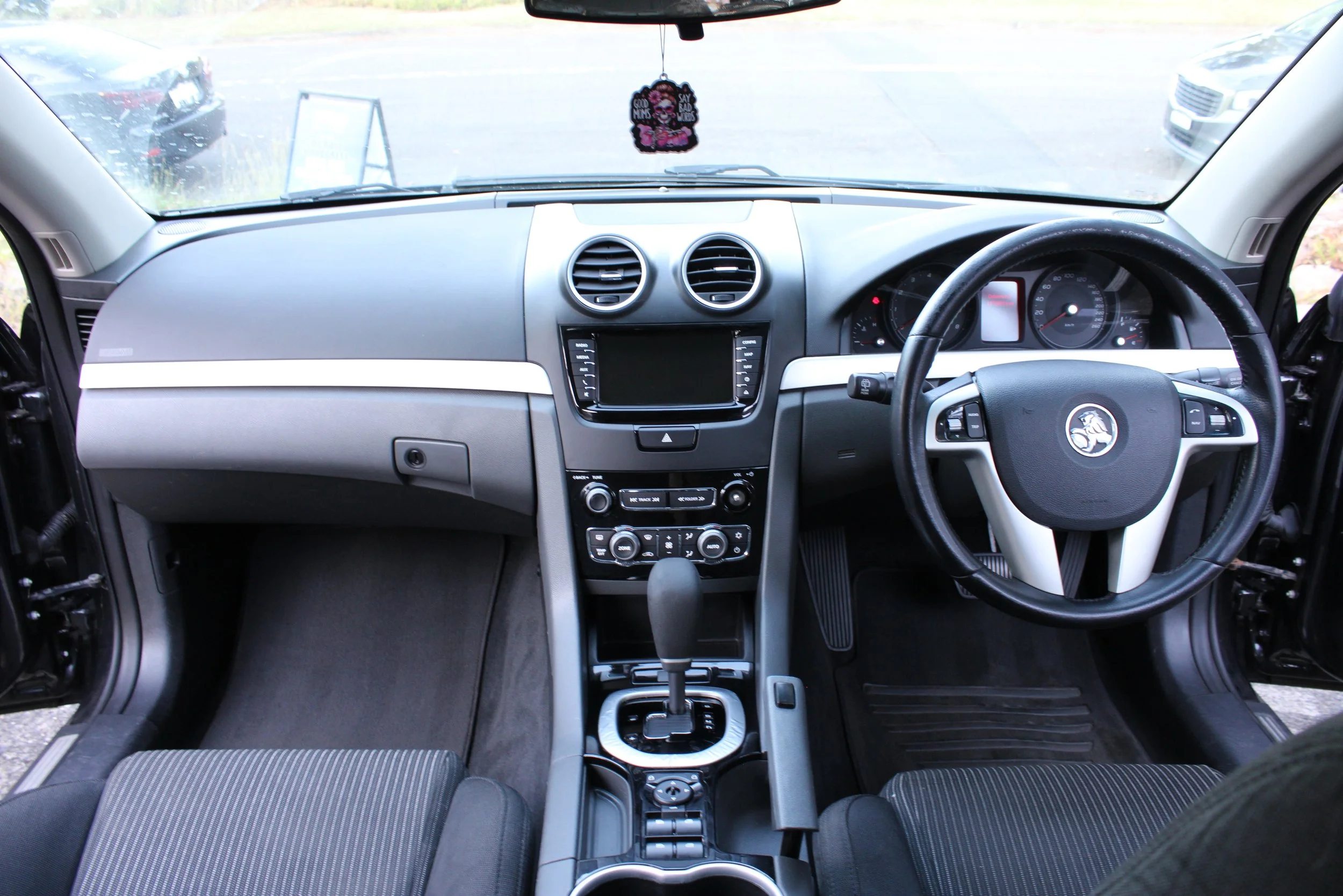 Interior of a vehicle dashboard showing the steering wheel on the right side, central console with touchscreen, air conditioning controls, gear shift, and driver's seat with visible seat fabric.