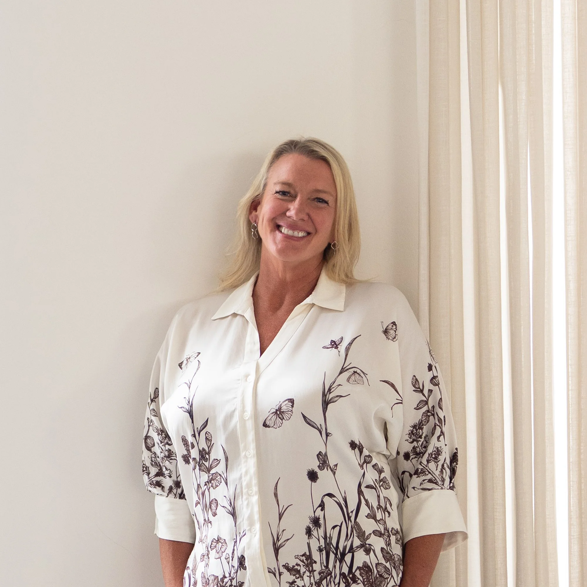 Interior designer Sally Wilson standing by sheer curtains in a light-filled space, wearing neutral tones and smiling warmly