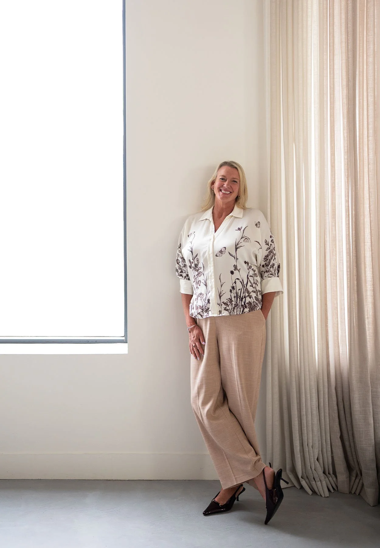 Interior designer Sally Wilson standing by sheer curtains in a light-filled space, wearing neutral tones and smiling warmly