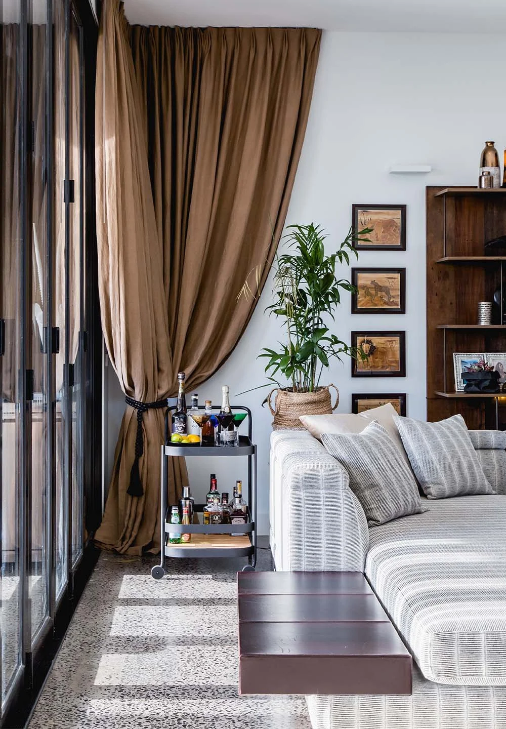 Living room corner with floor-length brown curtains, neutral sofa, styled bar cart and indoor plant creating a warm, layered interior setting