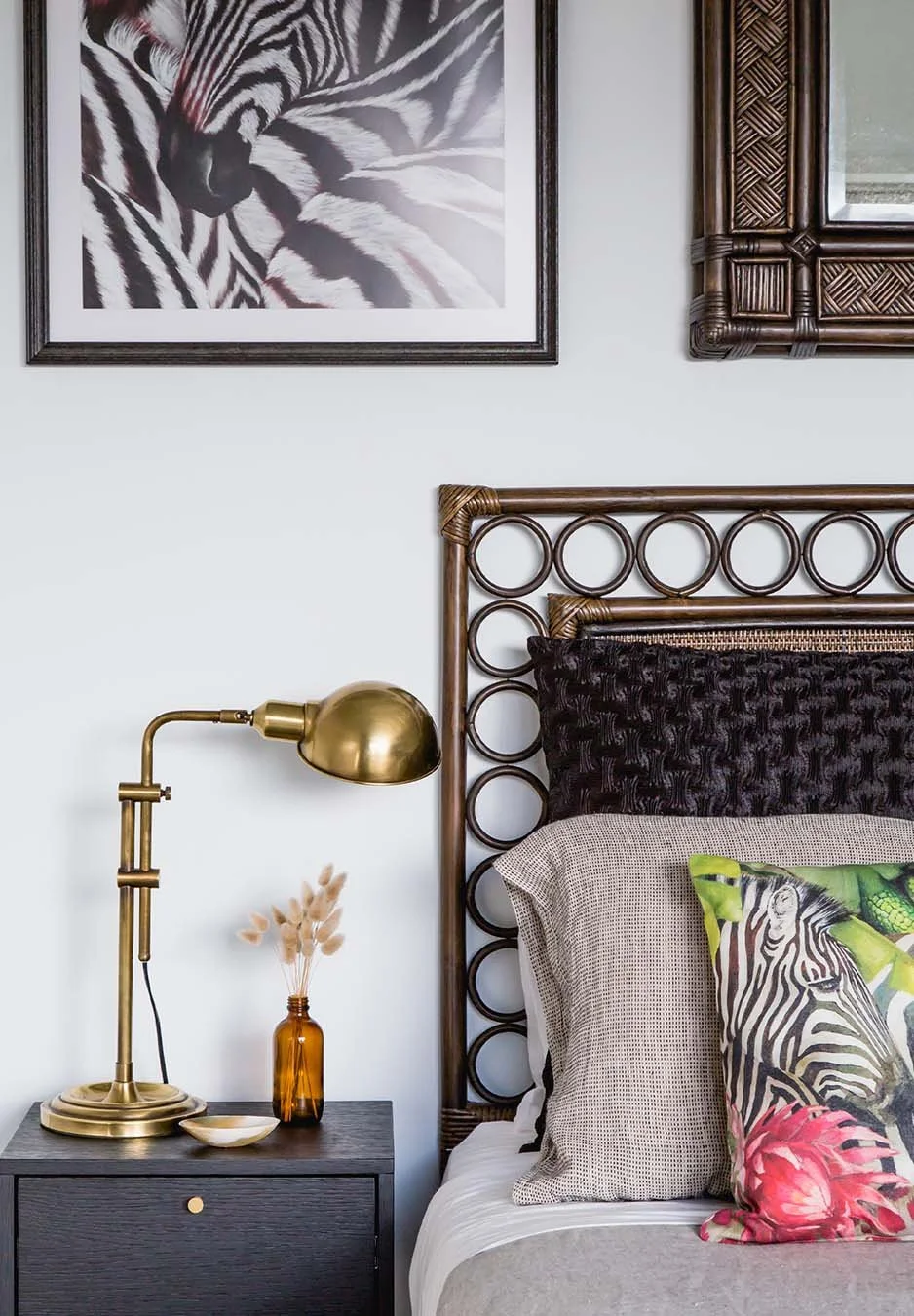 Bedroom detail with woven rattan headboard, brass reading lamp, textured cushions and zebra artwork creating a layered, curated bedside setting.