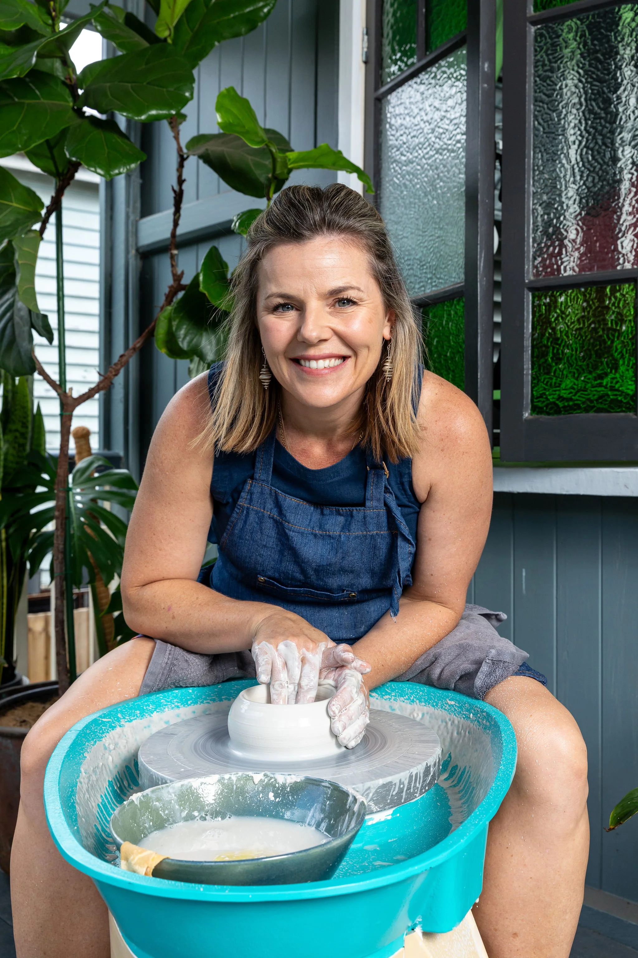 Woman smiling and working on a pottery wheel, shaping clay with her hands, surrounded by green plants in an undercover outdoor space.