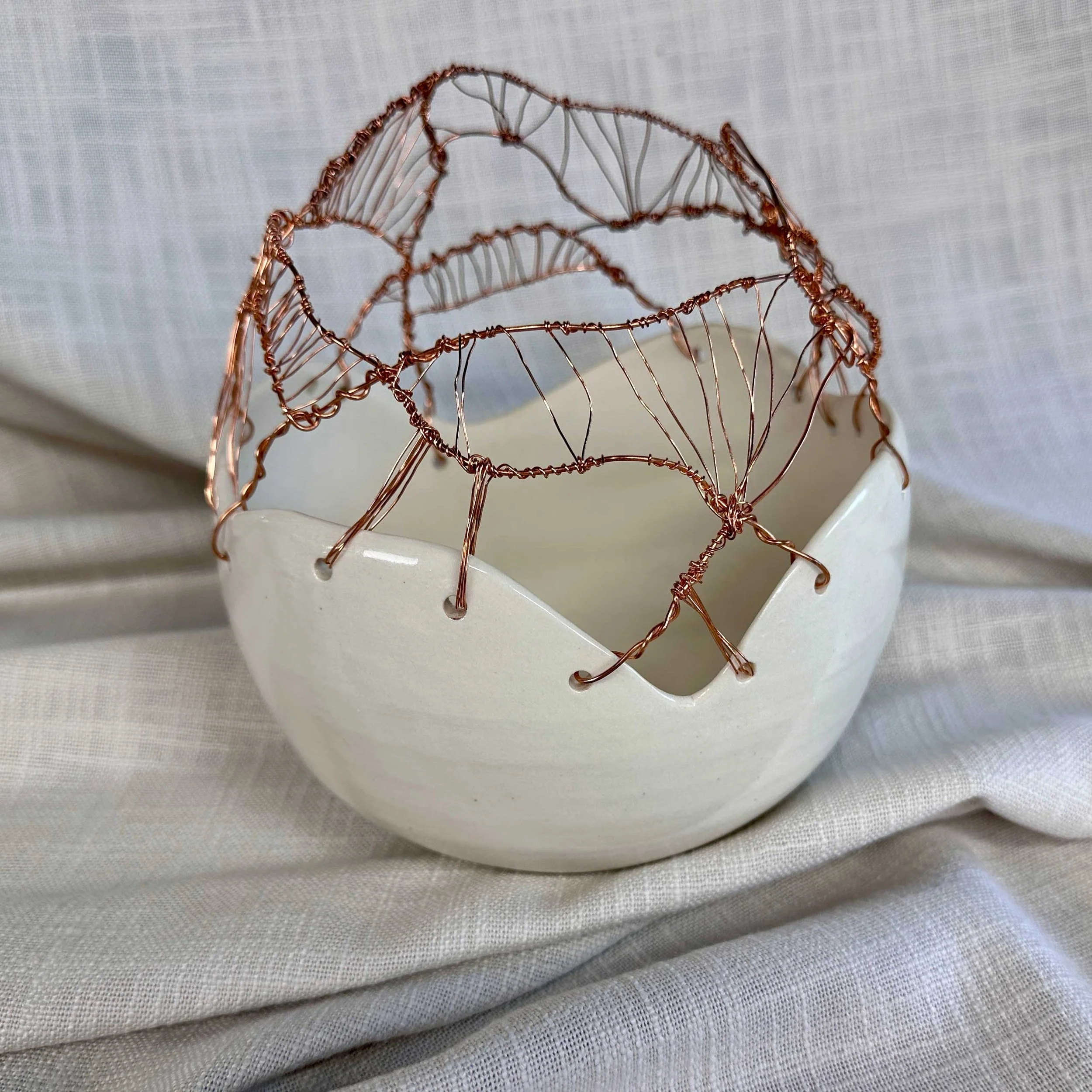 A white ceramic bowl with a jagged edge, decorated with a delicate wire sculpture resembling a butterfly, made from copper-colored wire, placed on light-colored fabric