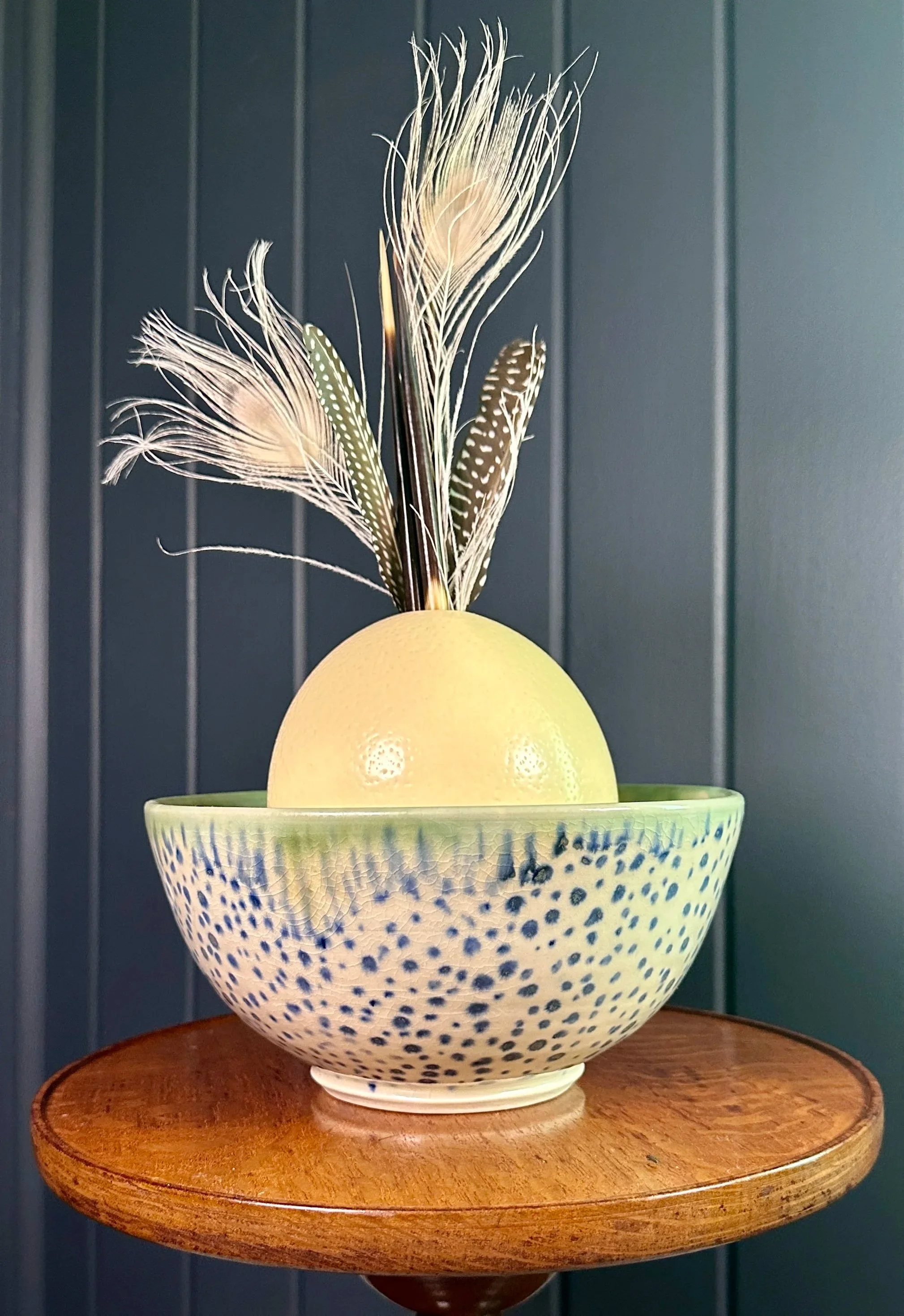 Decorative bowl with an ostrich egg and peacock feathers on a wooden table against a dark vertical panel wall.