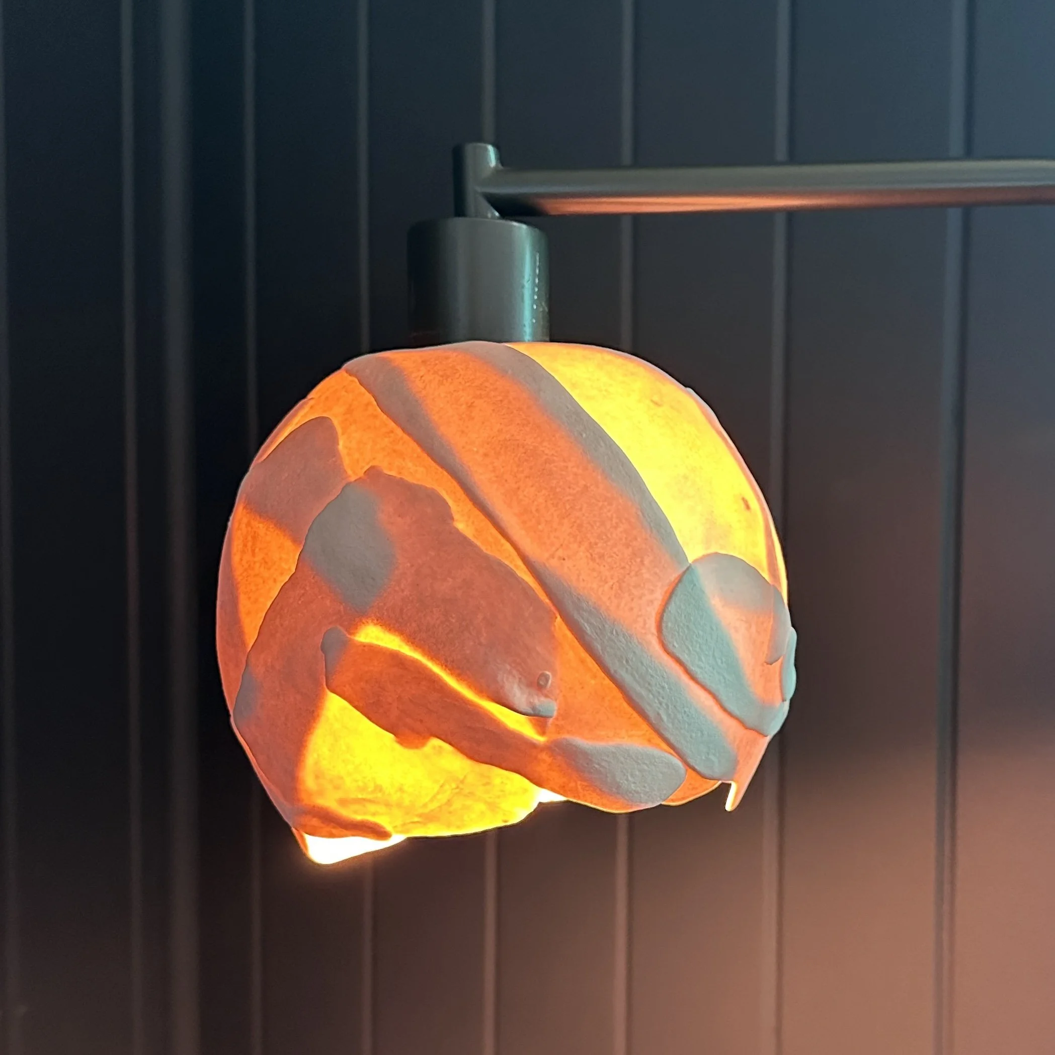 A handmade porcelain lamp with abstract design glowing orange through the translucent material, set against a dark charcoal wall. 