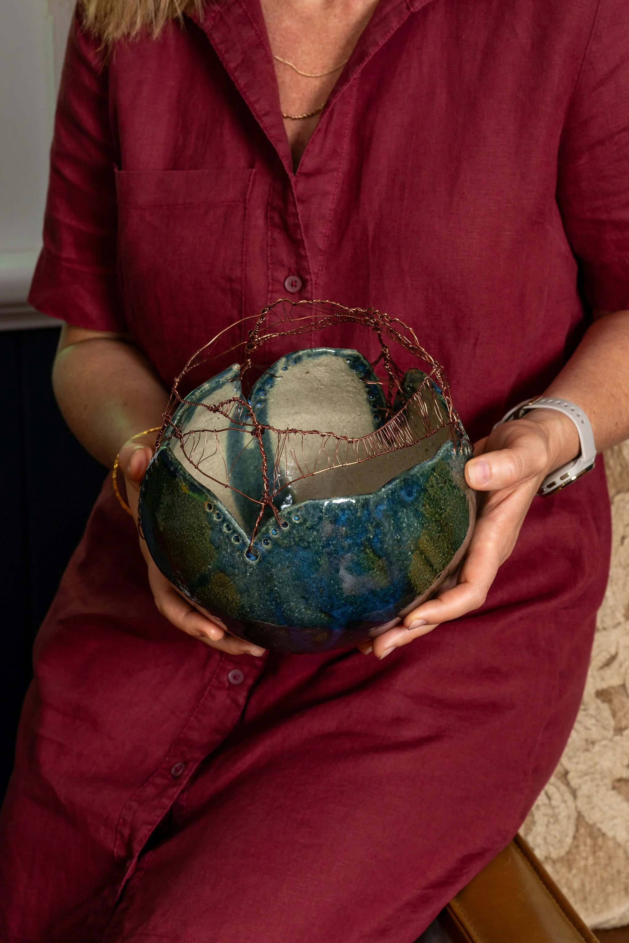 A person wearing a red shirt holds a ceramic lampshade with a textured, colorful glaze and an intricate copper wire structure on top.