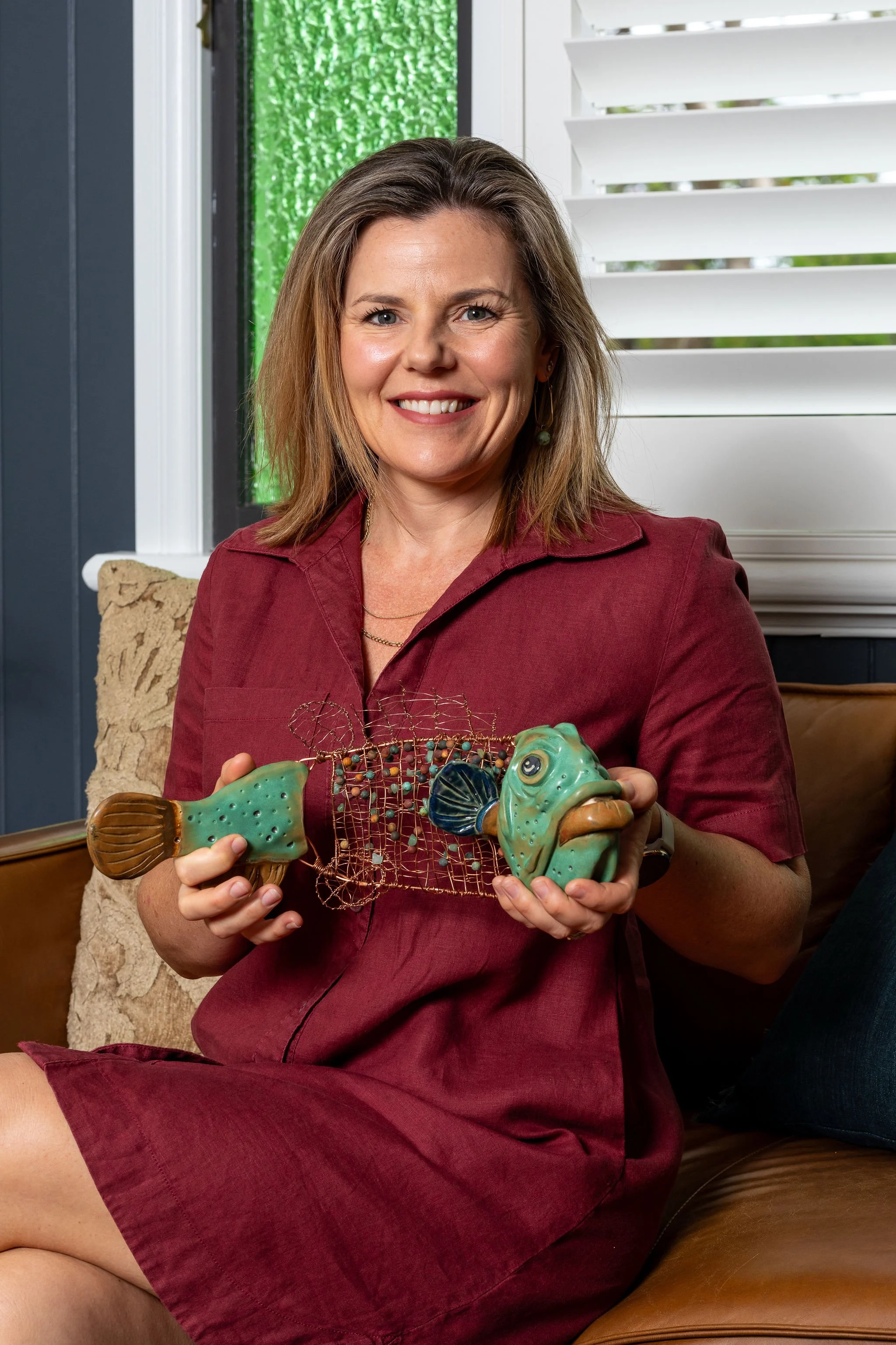 A woman with shoulder-length brown hair smiling and sitting on a leather couch, holding a ceramic fish sculpture in her hands, in a room with a window and white blinds.