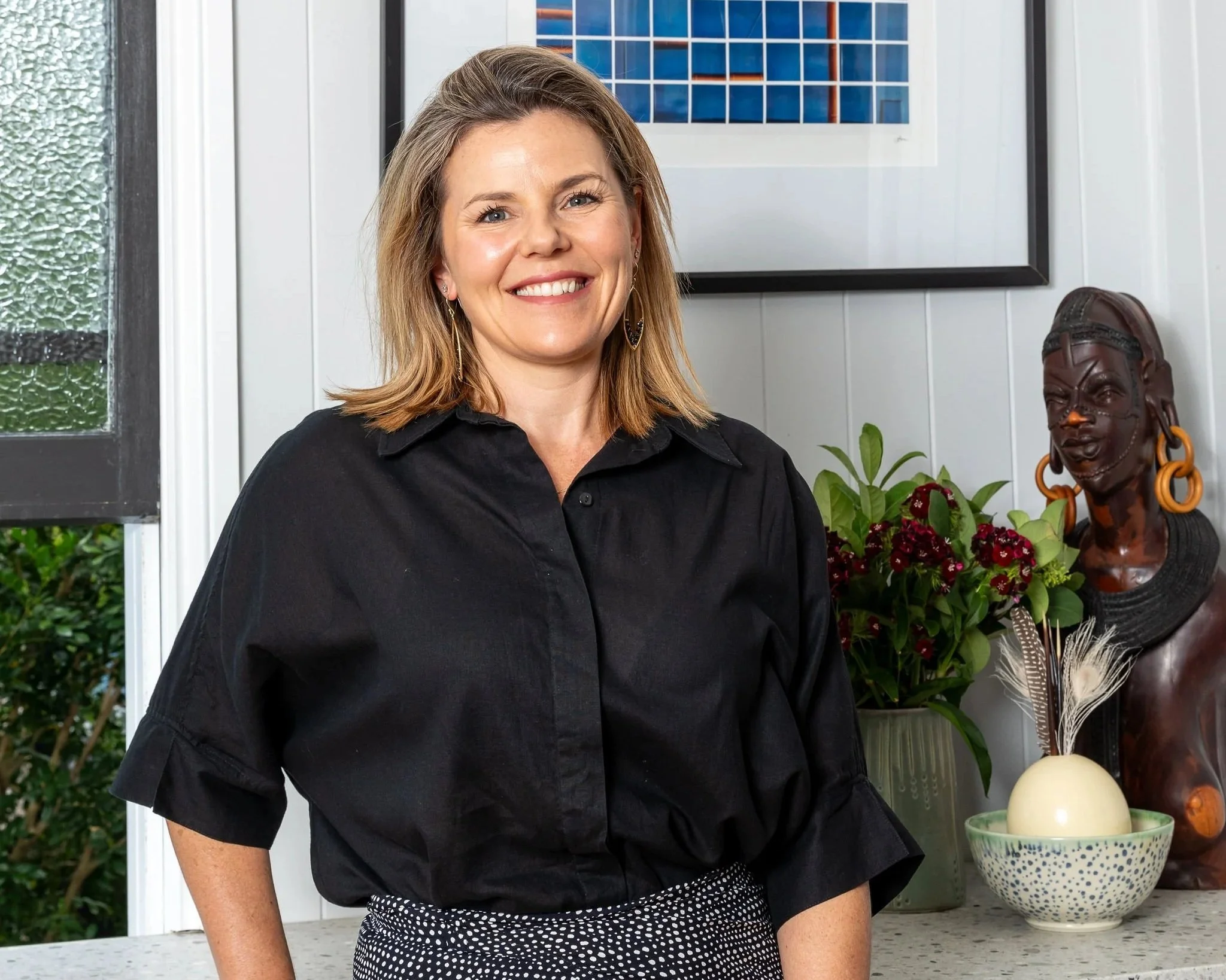 Smiling woman with shoulder-length light brown hair wearing a black shirt standing in a bright room with a decorative sculpture, potted plant, and framed artwork in the background.