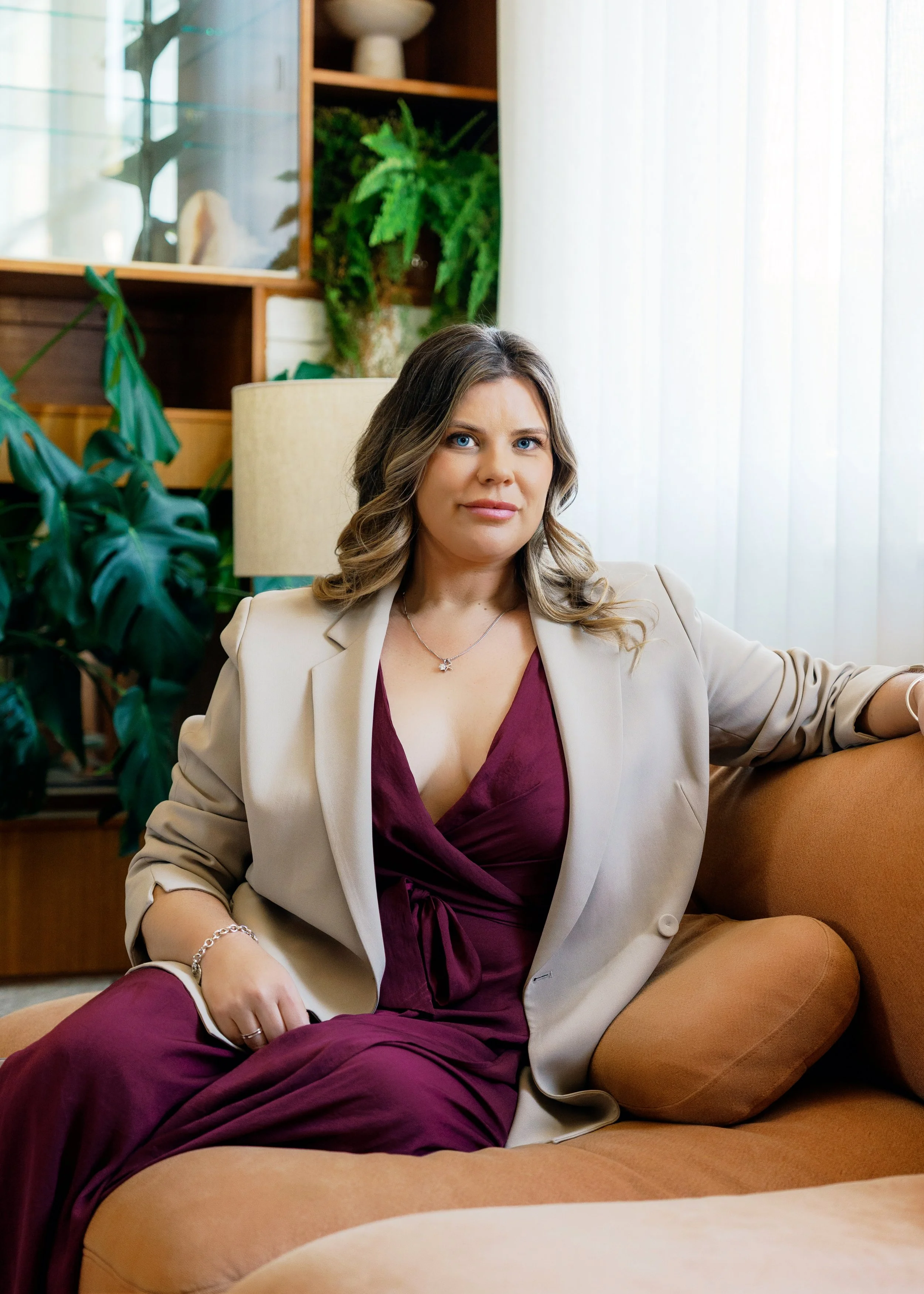 Bidpop founder, Jessica Tunnicliff, seated confidently in a warm, plant-filled interior, wearing a burgundy wrap dress and tailored beige blazer.