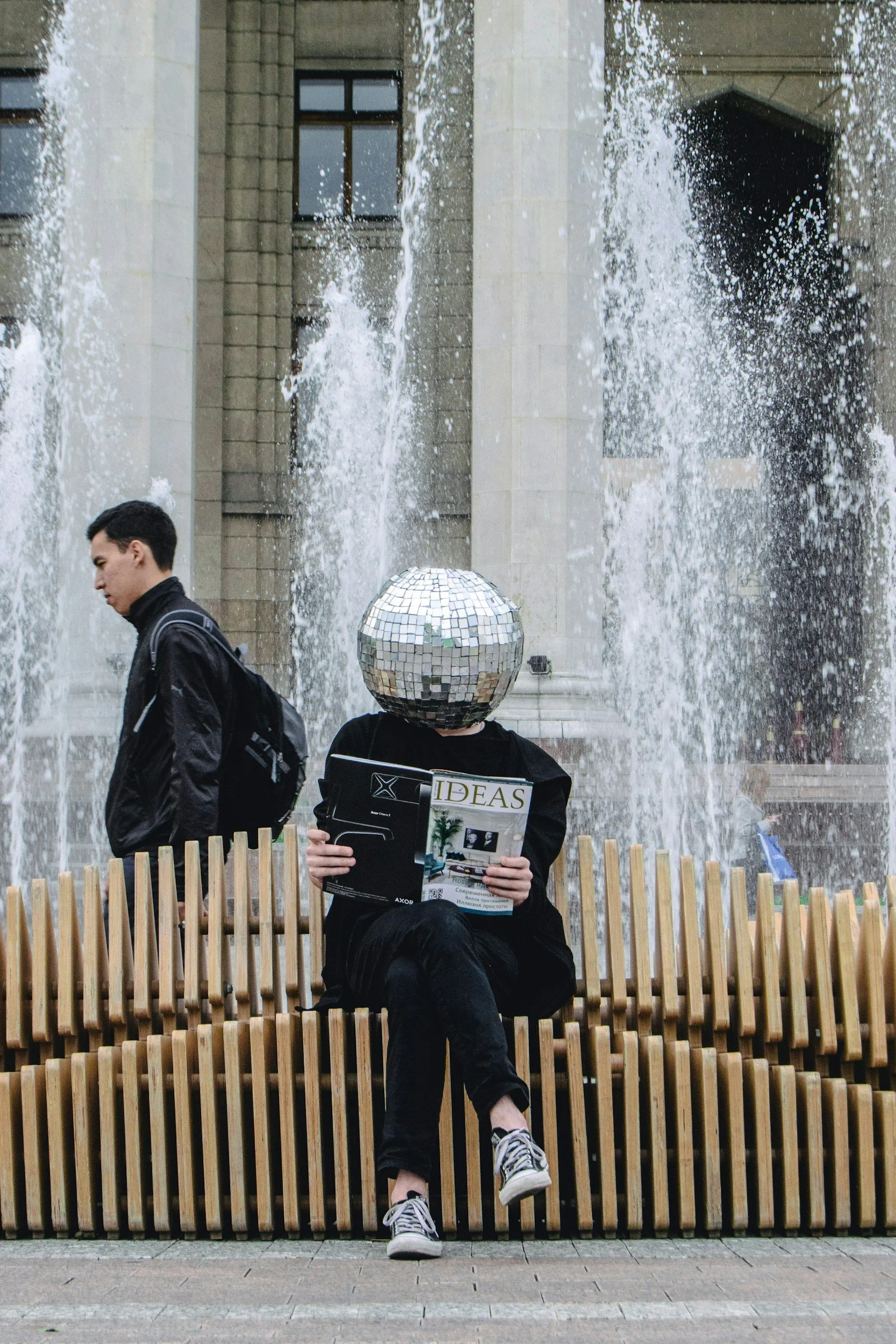 Person sitting on a wooden bench reading a magazine with a disco ball as a head, in front of a fountain with water sprays in an urban plaza, while another person walks by